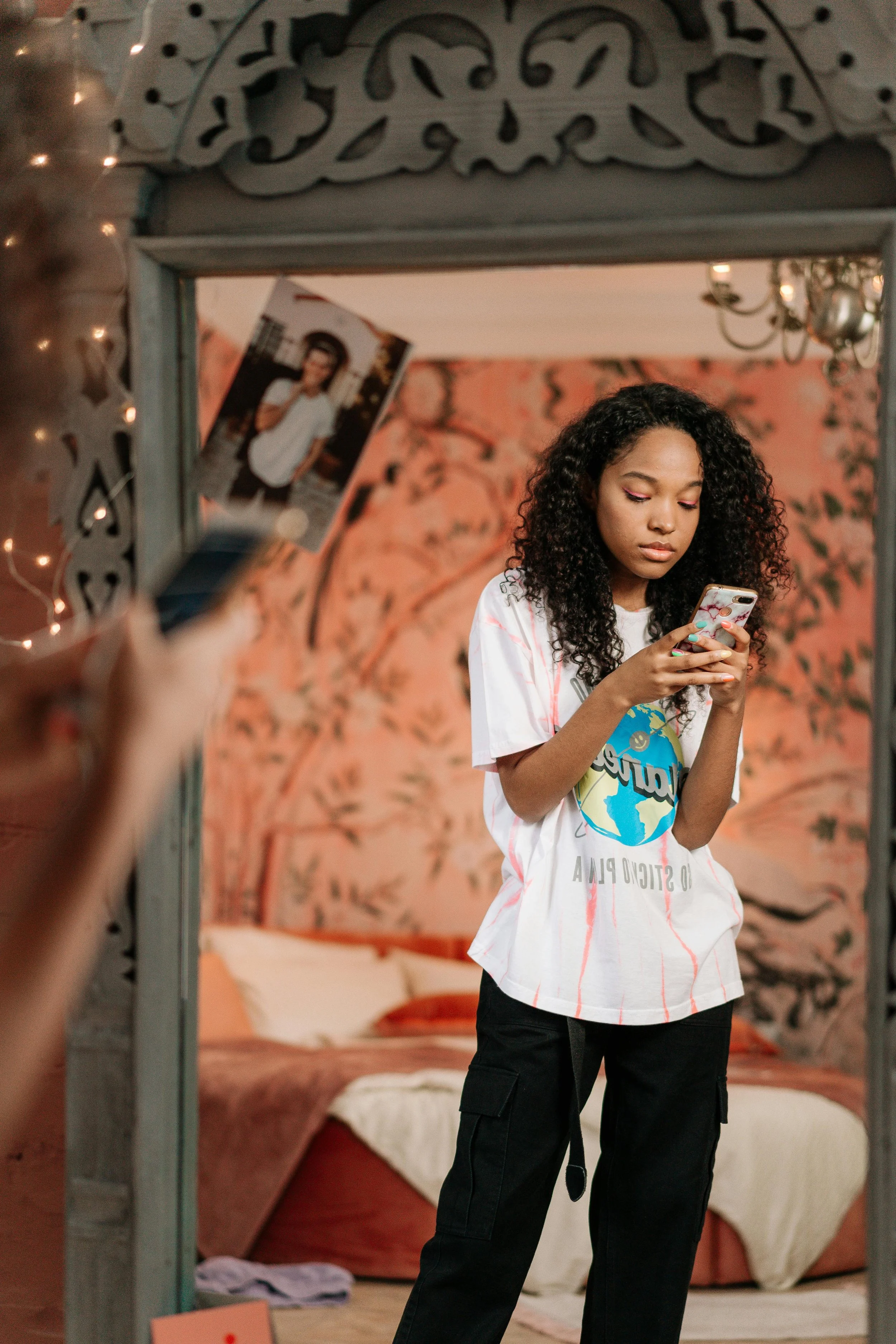 A young woman with curly hair looking at her phone, standing in front of a mirror in a cozy room with a bed and patterned wallpaper.