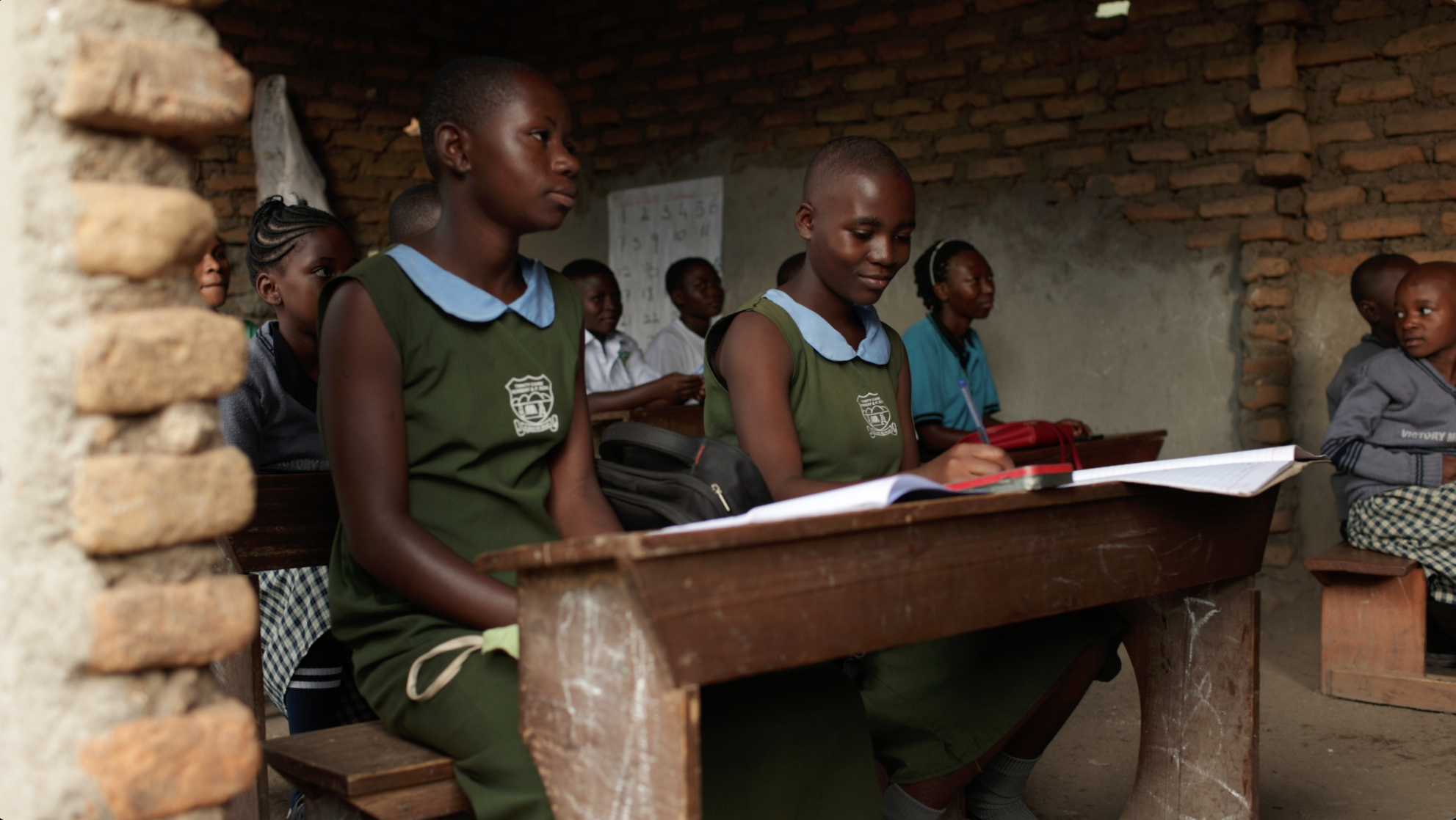 Students sitting in a rustic classroom with brick walls, one writing in a notebook while others look on.