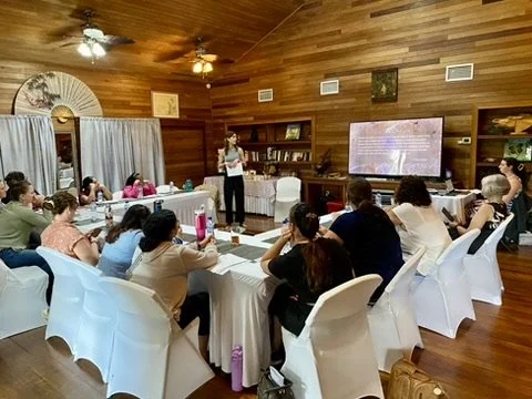 A woman giving a presentation in a room with wooden walls and ceiling, audience seated at tables, large screen displaying a slide, bookshelves and decorative items visible.