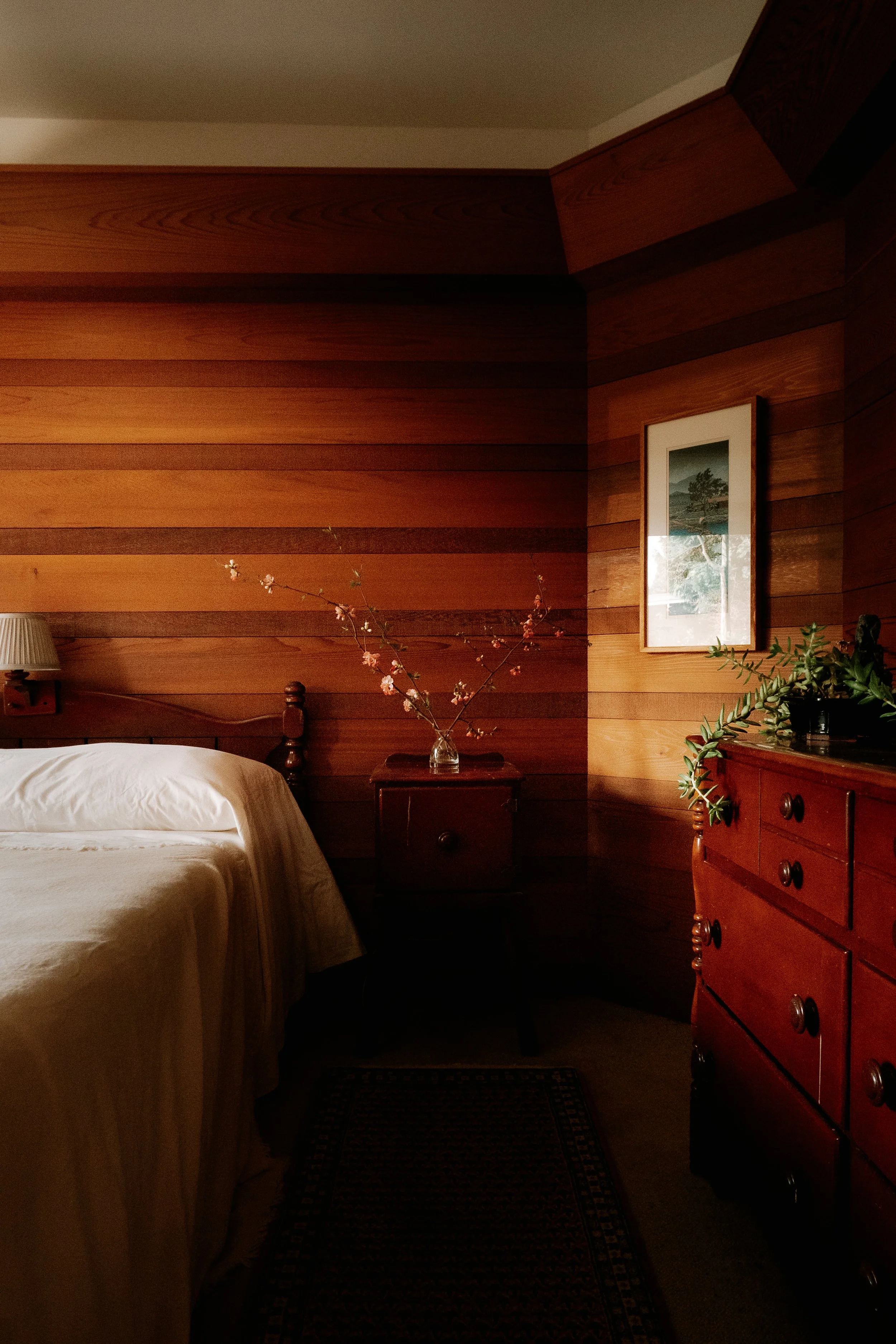 A bedroom corner with wooden paneled walls, a bed with white bedding, a bedside table with a vase of pink flowers, a framed picture, and a wooden dresser with a plant
