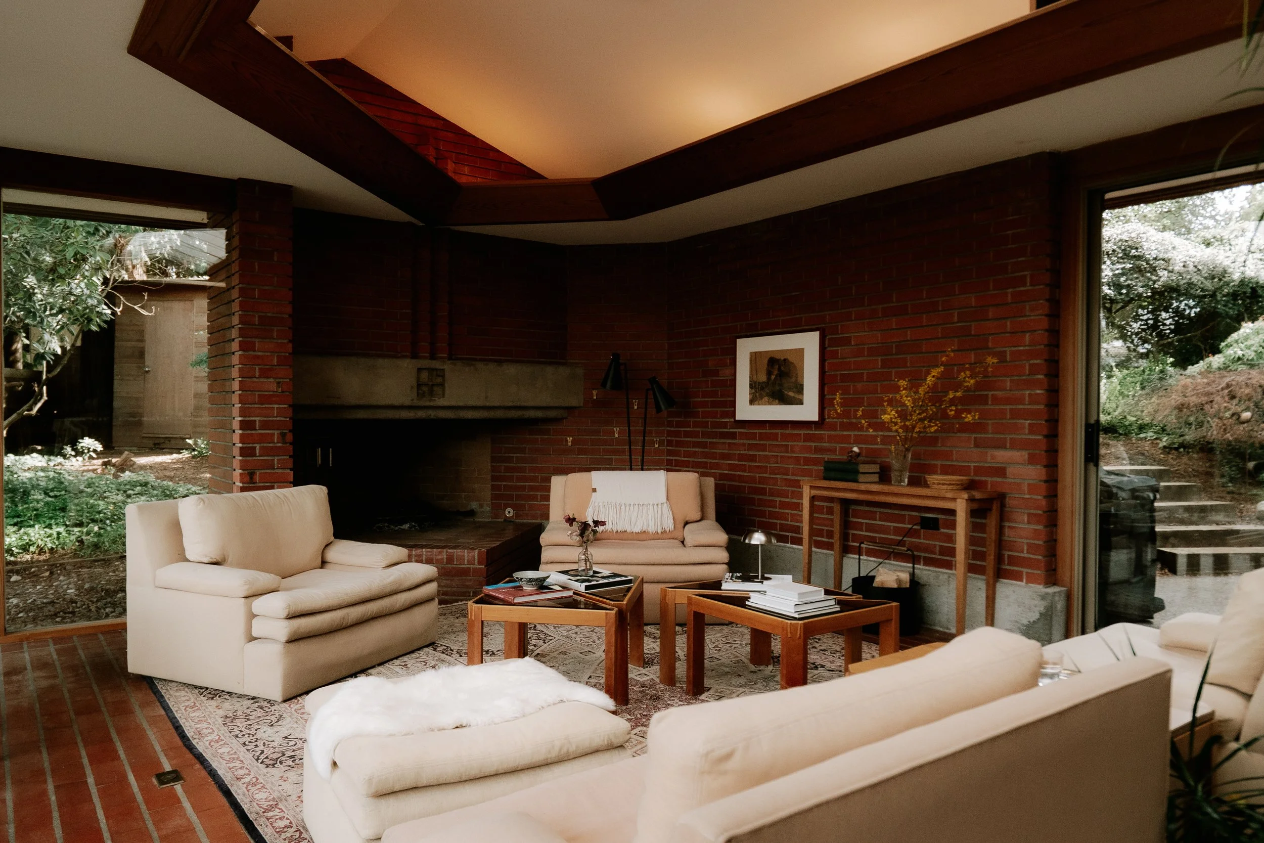 Interior of a living room with beige sofas, a coffee table with books, and a brick wall with a fireplace. Large windows show a garden outside. The ceiling has wooden beams, and decor includes framed artwork and a vase with yellow flowers.