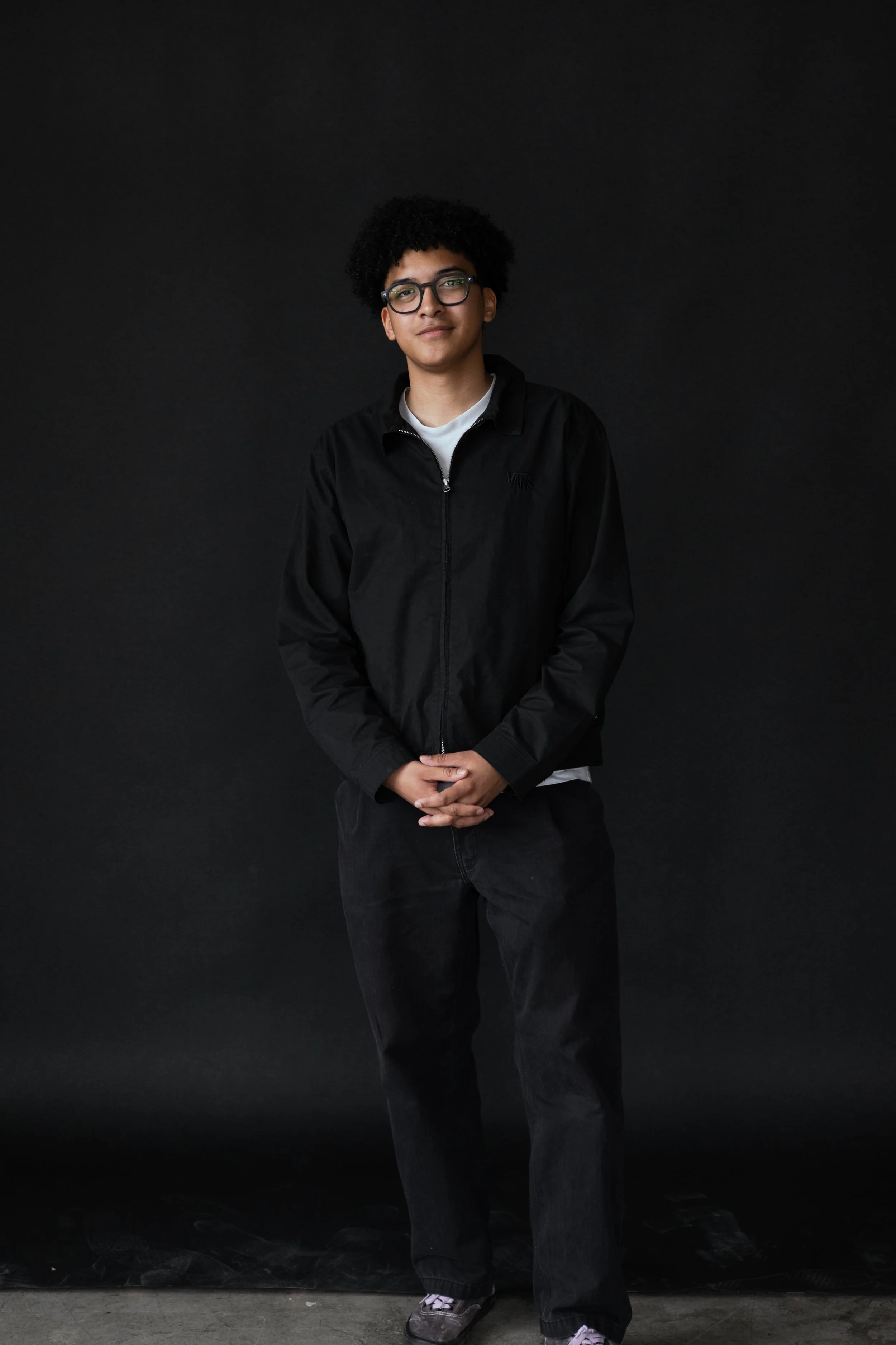 Young man with curly hair and glasses standing against a black background, wearing a black jacket and dark pants.