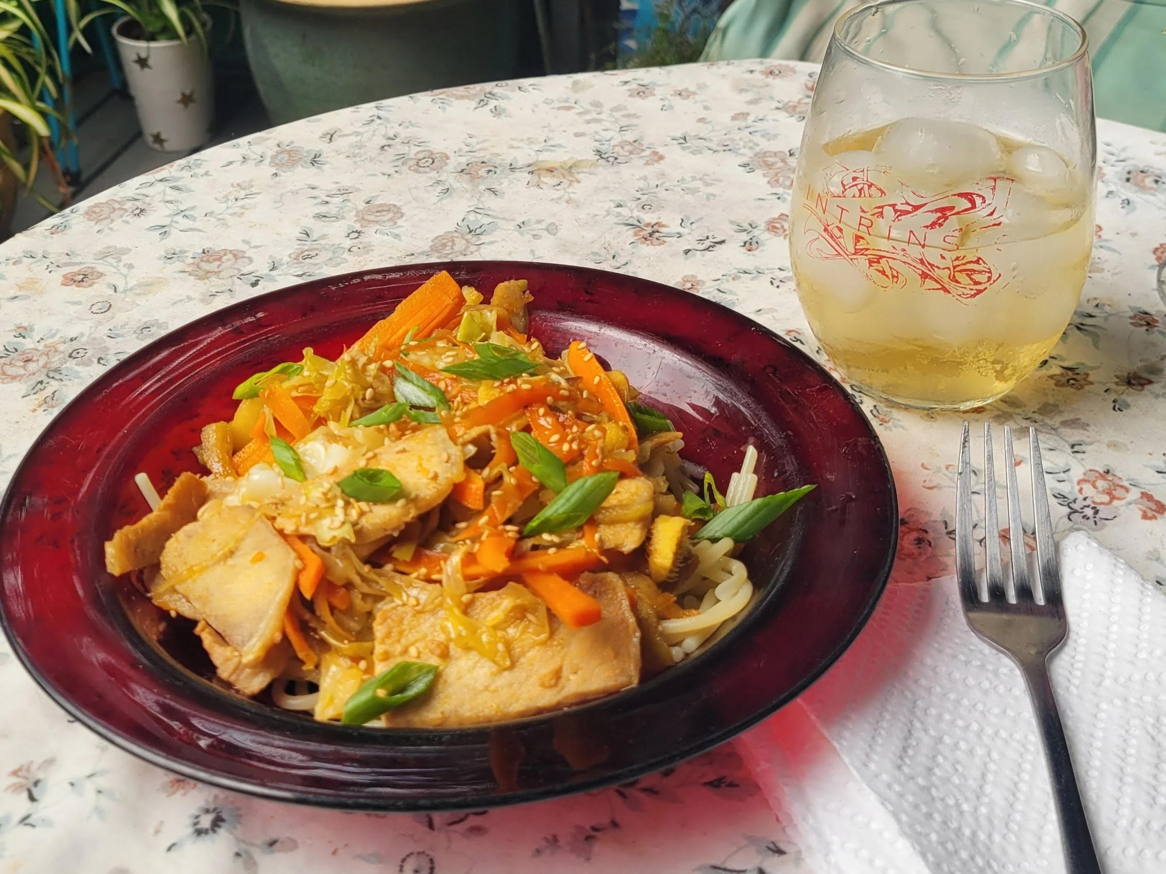 Ginger pork and cabbage stir fry with orange ginger sauce served on a flowered tablecloth outdoors, with a fork, napkin, drink, and garden visible in the background.