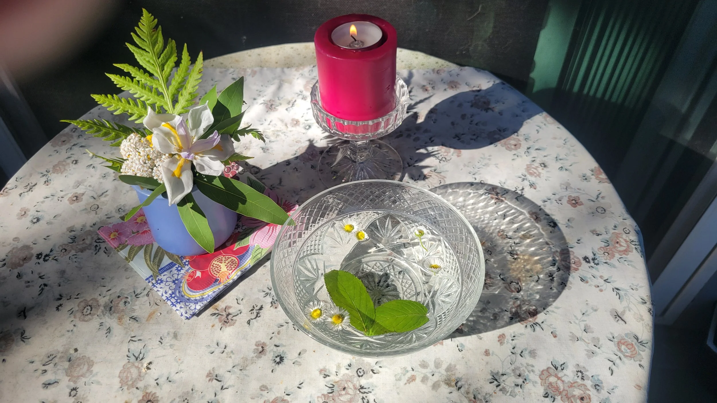 A small patio table with a floral tablecloth, fairy bouquet, red candle, and crystal bowl filled with floating daisies and peppermint leaves