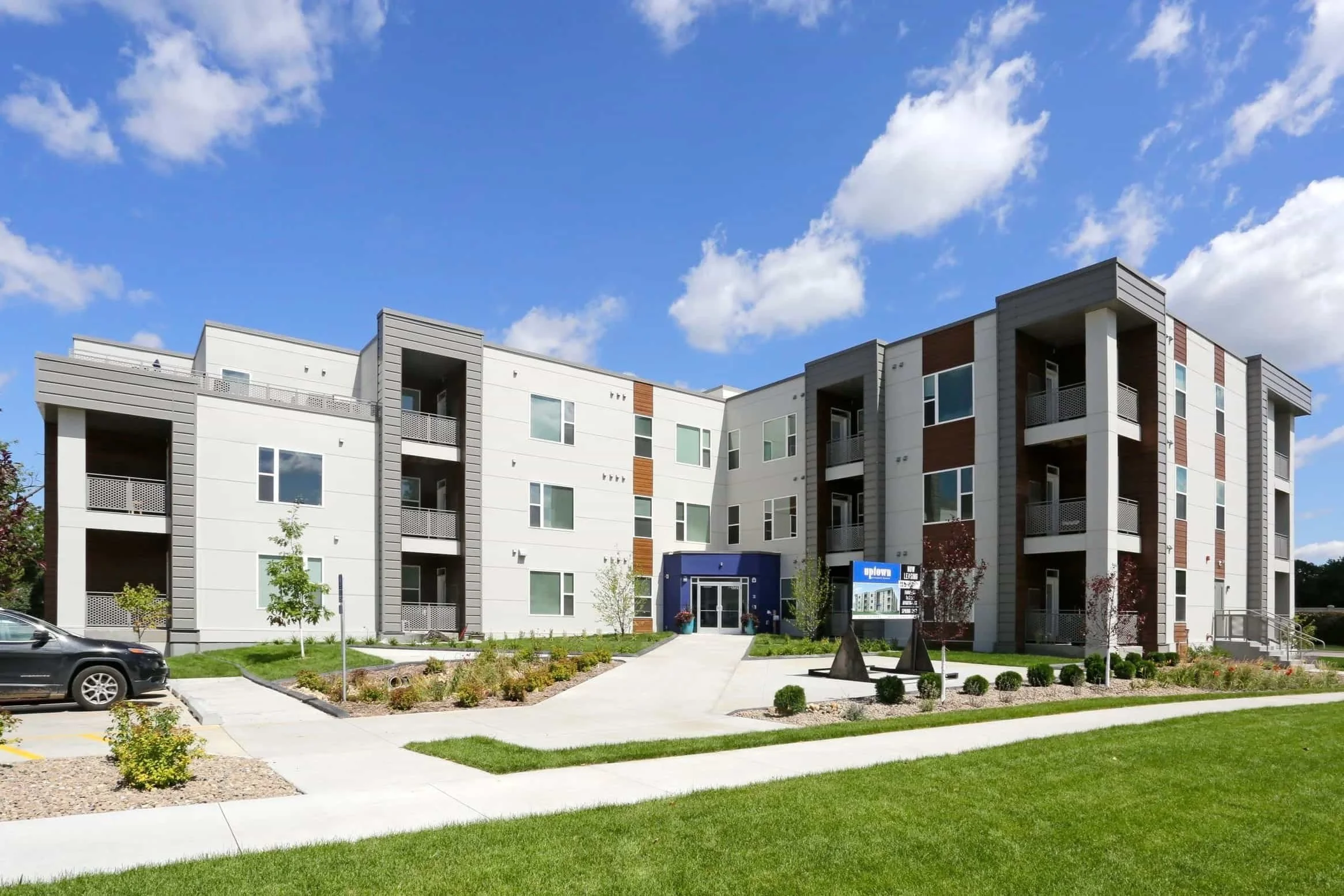 Modern three-story apartments in Rochester with balconies, landscaped lawn, and a blue sky with scattered clouds.