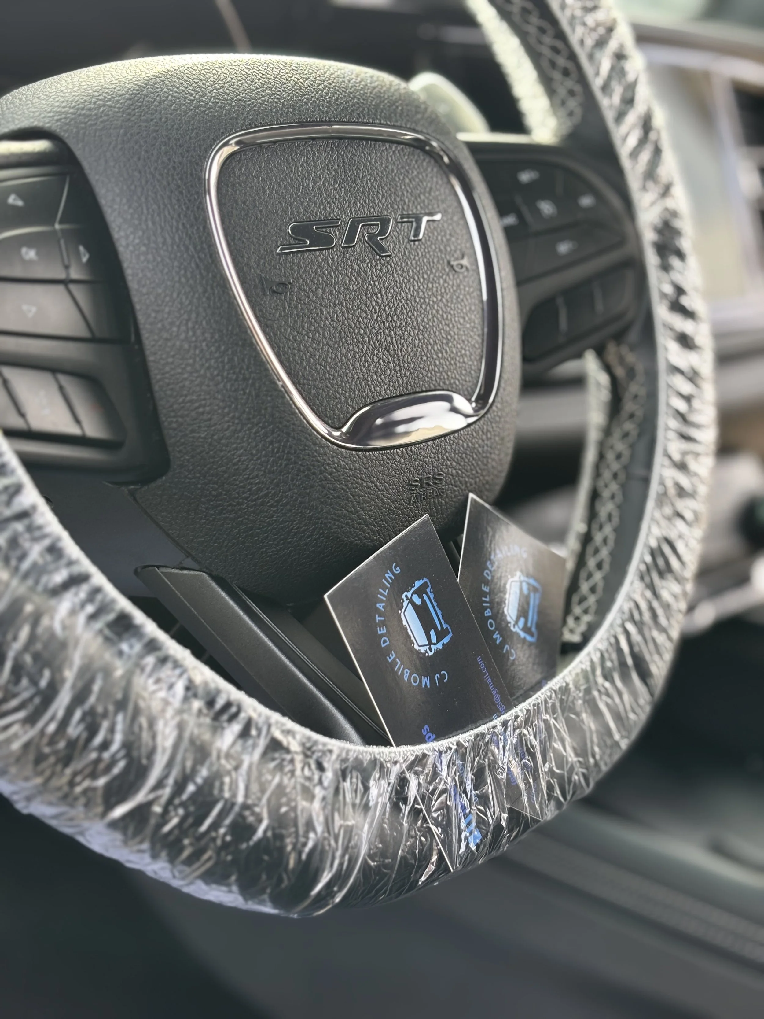 Close-up of a Dodge SRT steering wheel covered with a clear plastic wrap, with two business cards or cards with logo and text in the center of the wheel.
