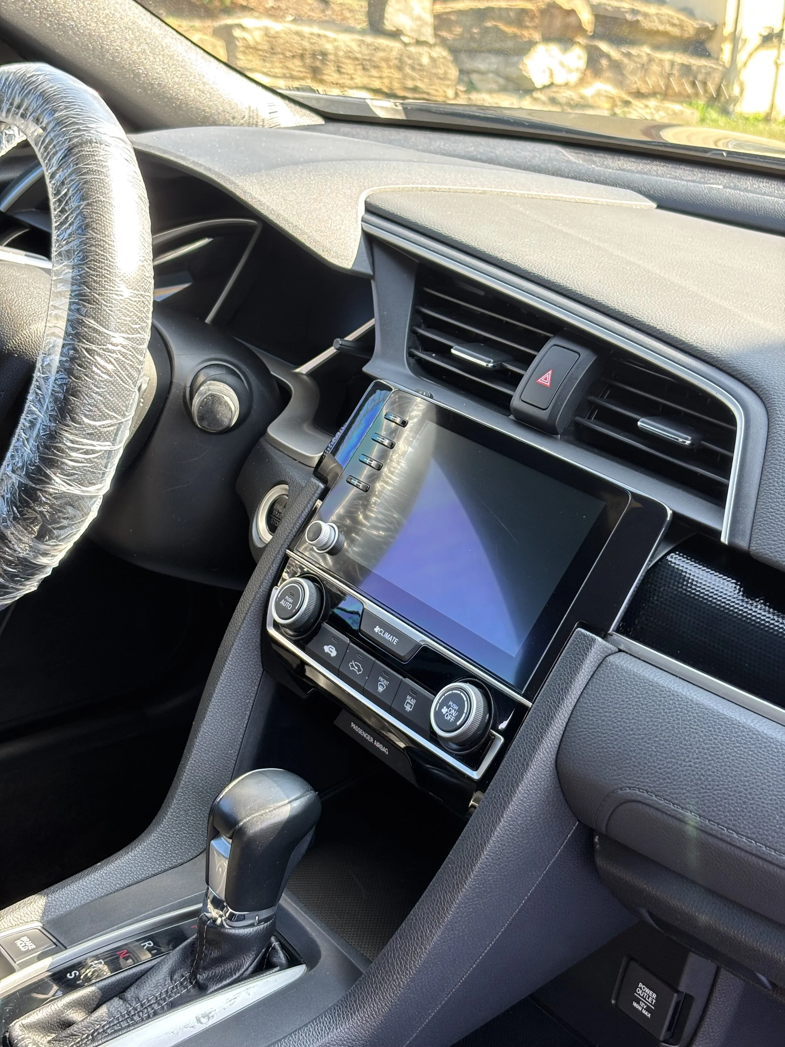 Interior of a modern car showing the dashboard, touchscreen display, climate controls, gear shift, and steering wheel with a plastic covering.