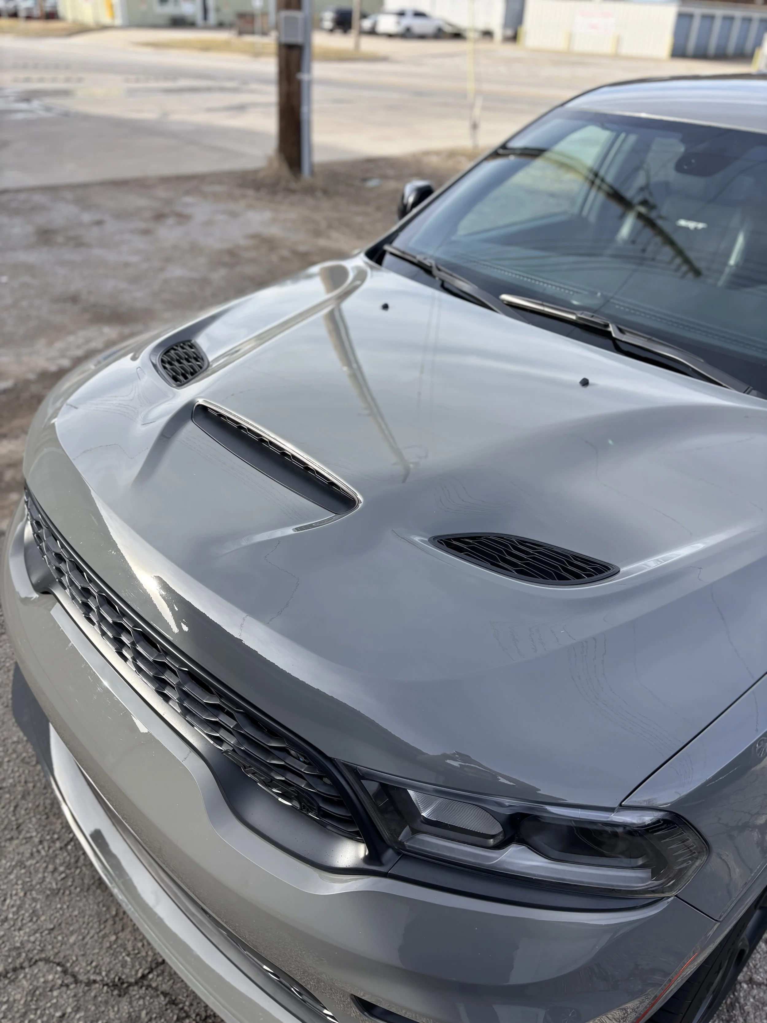 Front of a silver car with hood vents, parked on a gravel lot near a street and industrial buildings.