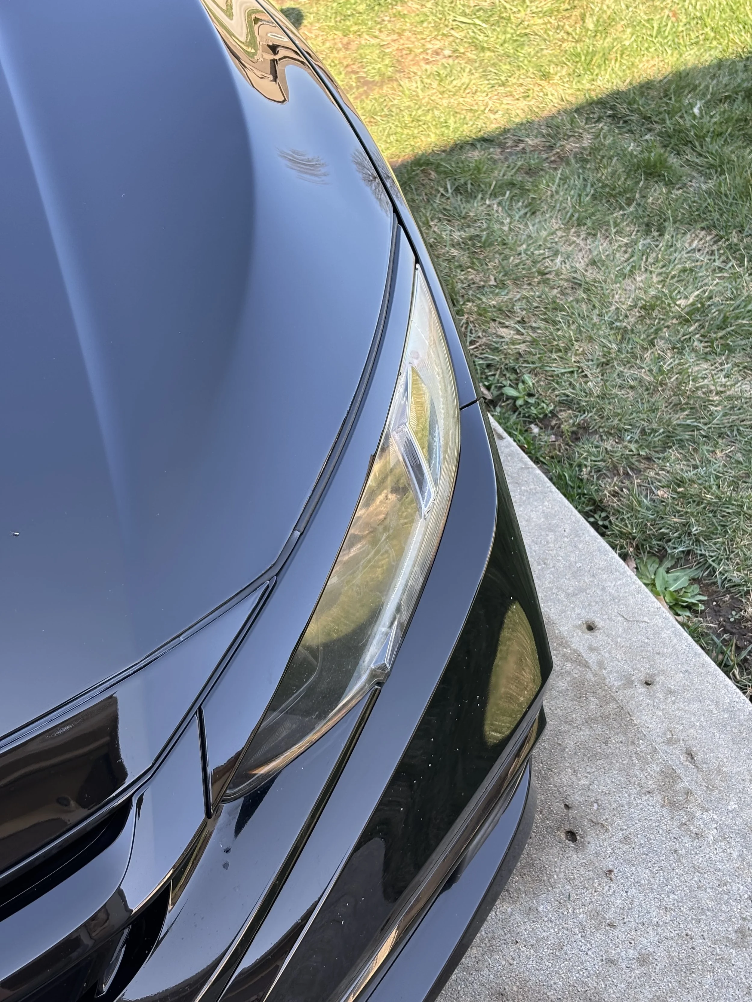 Close-up view of the front end of a black car parked on a concrete driveway, with grass and soil visible nearby.