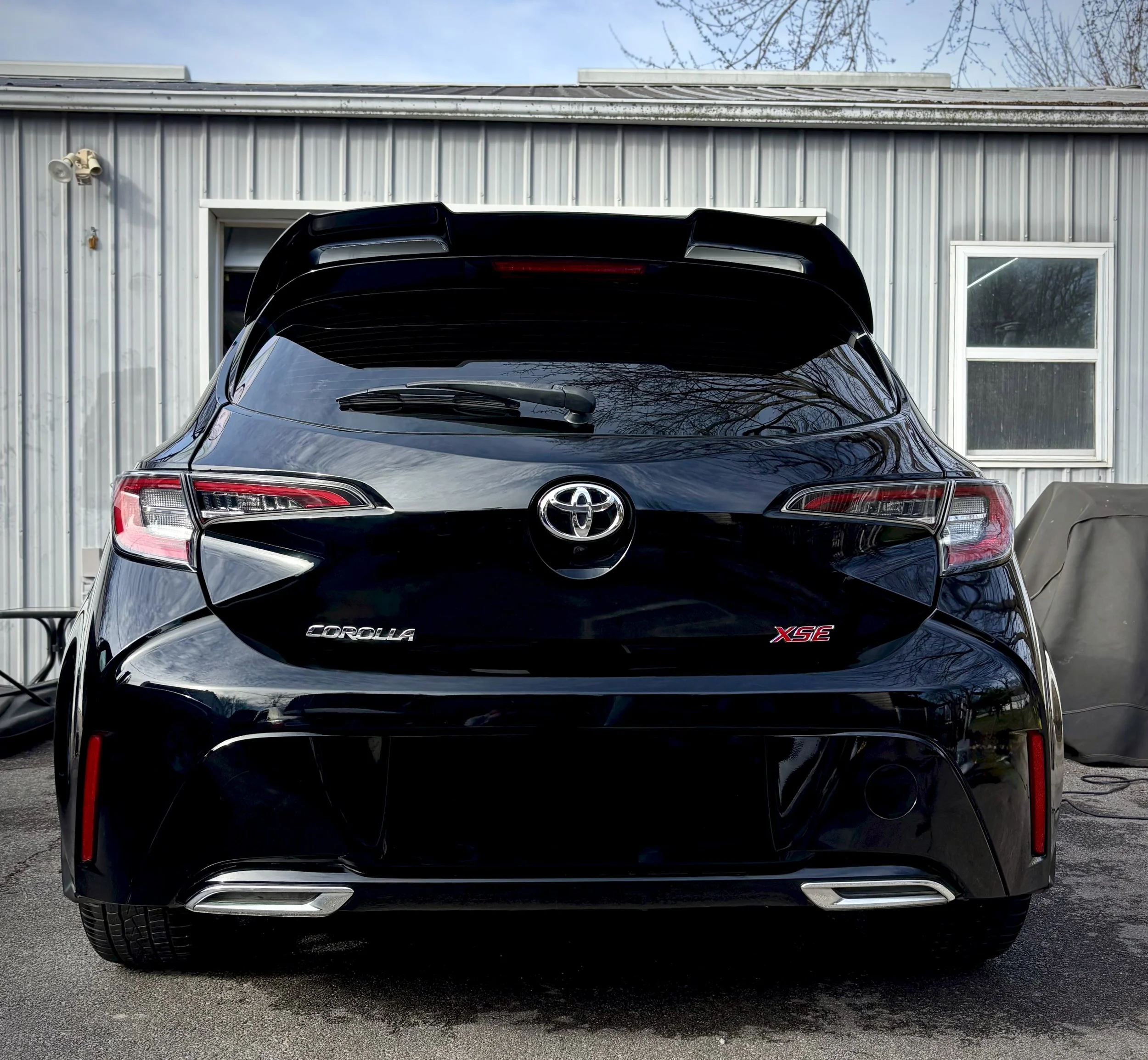 Black Toyota Corolla XSE parked outside a building with metal siding and a window, with reflections of trees on the car's surface.