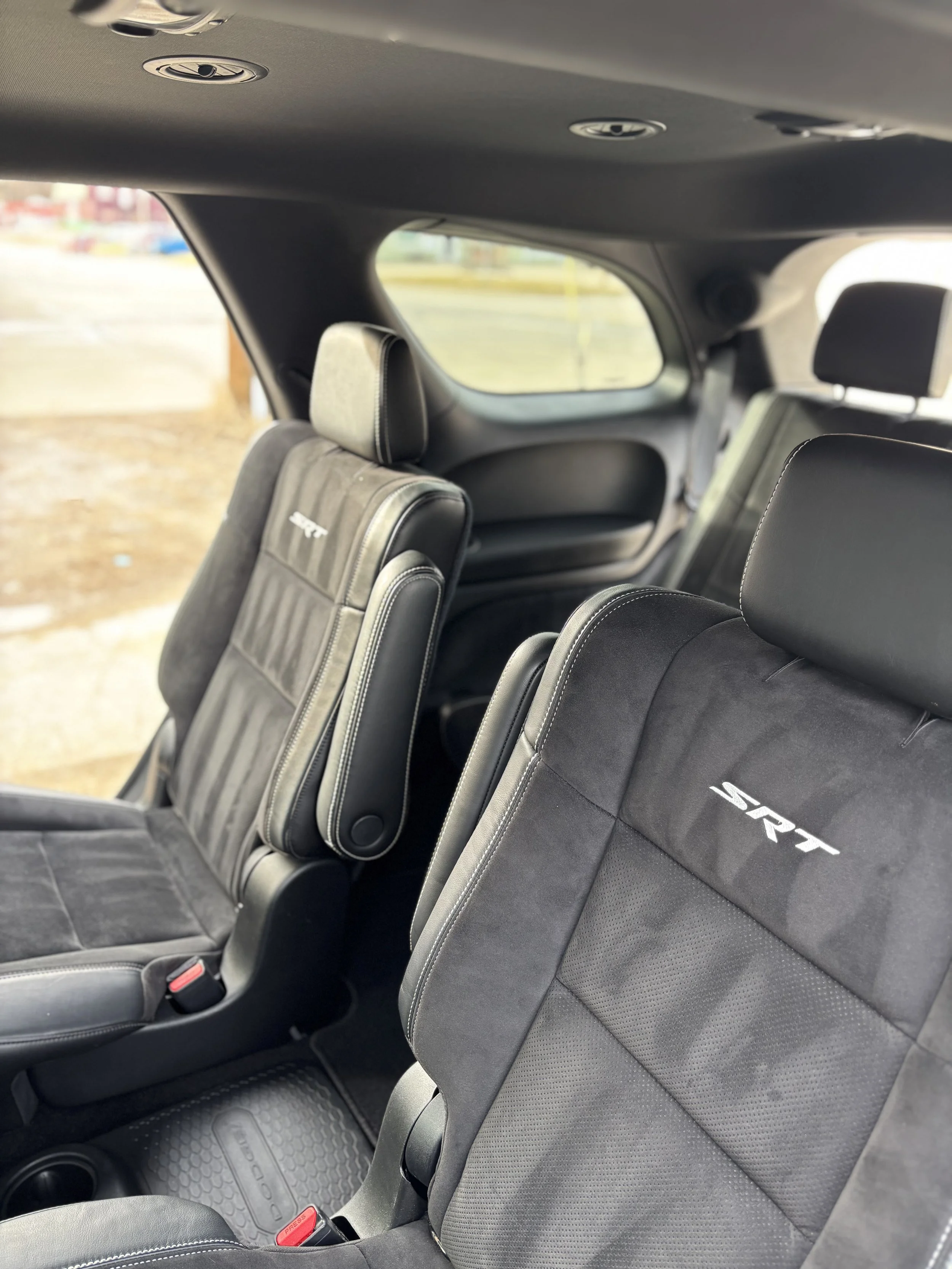 Interior of a vehicle showing black seats with 'SRT' embroidery, a fold-up seat in the middle, and a large window in the background.