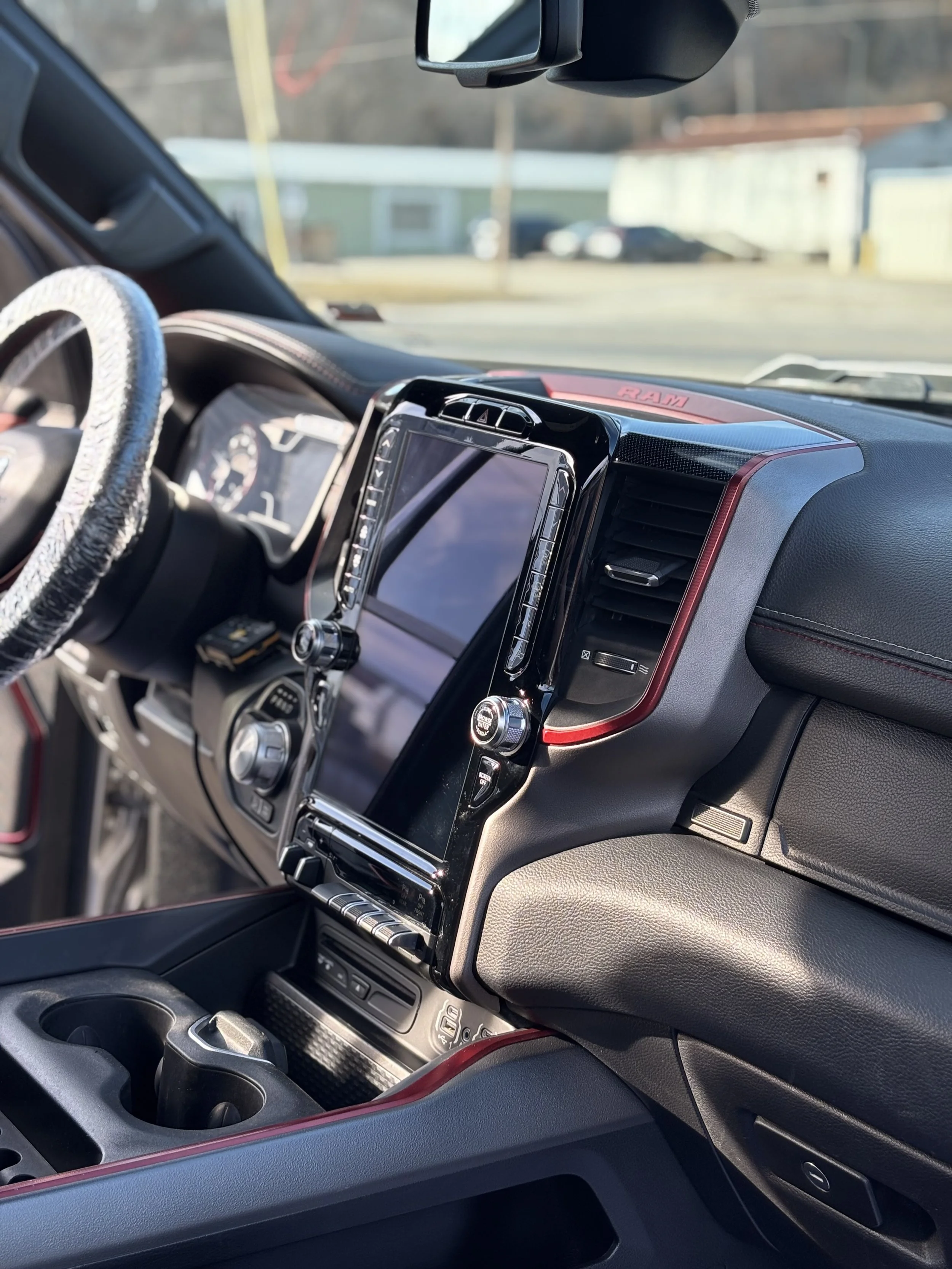 The interior of a truck's dashboard, featuring a large touchscreen display, climate controls, cup holders, and a steering wheel wrapped in a textured cover. Outside the windshield, a blurry background of buildings and cars can be seen.
