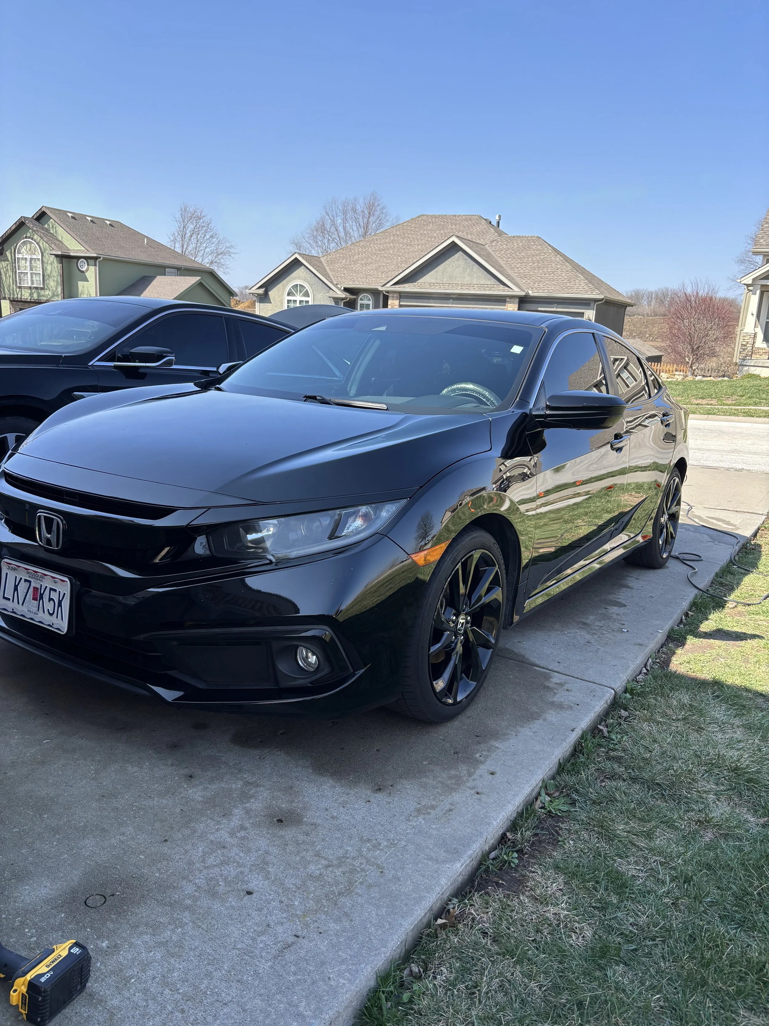 A black Honda sedan parked on a concrete driveway in front of a suburban house, with another car partially visible beside it, and residential homes in the background under a clear blue sky.