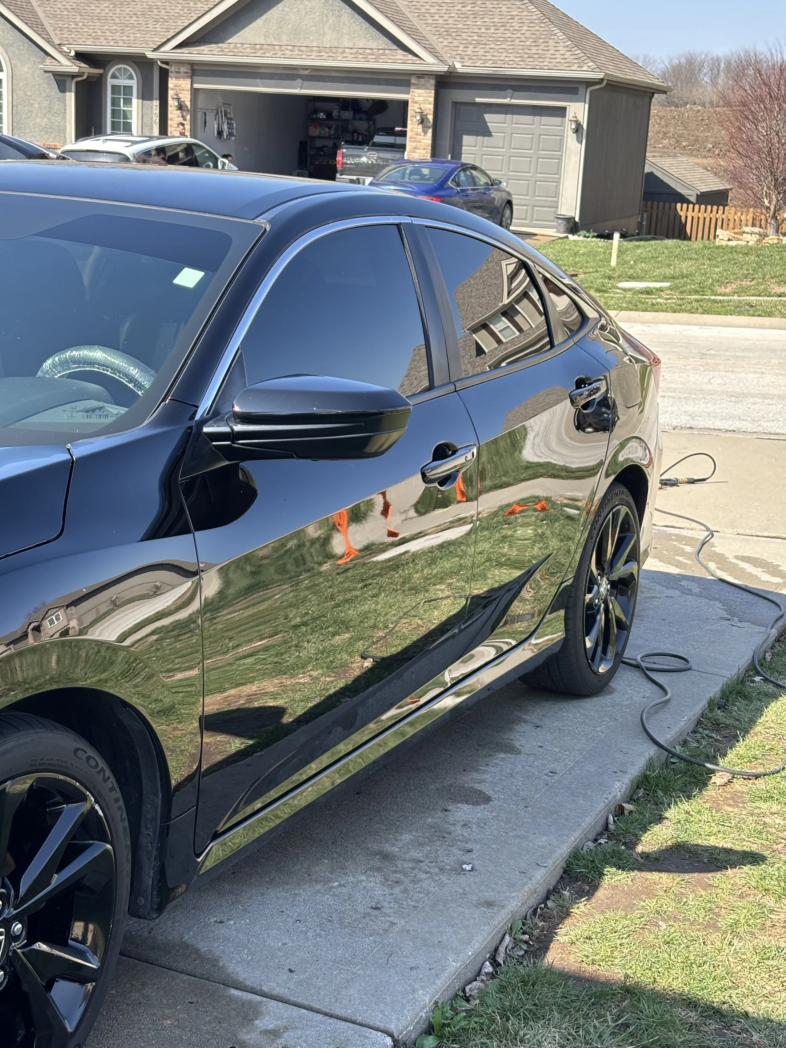 A black sedan car parked on a concrete driveway, connected to an outdoor power source, with a house garage in the background.