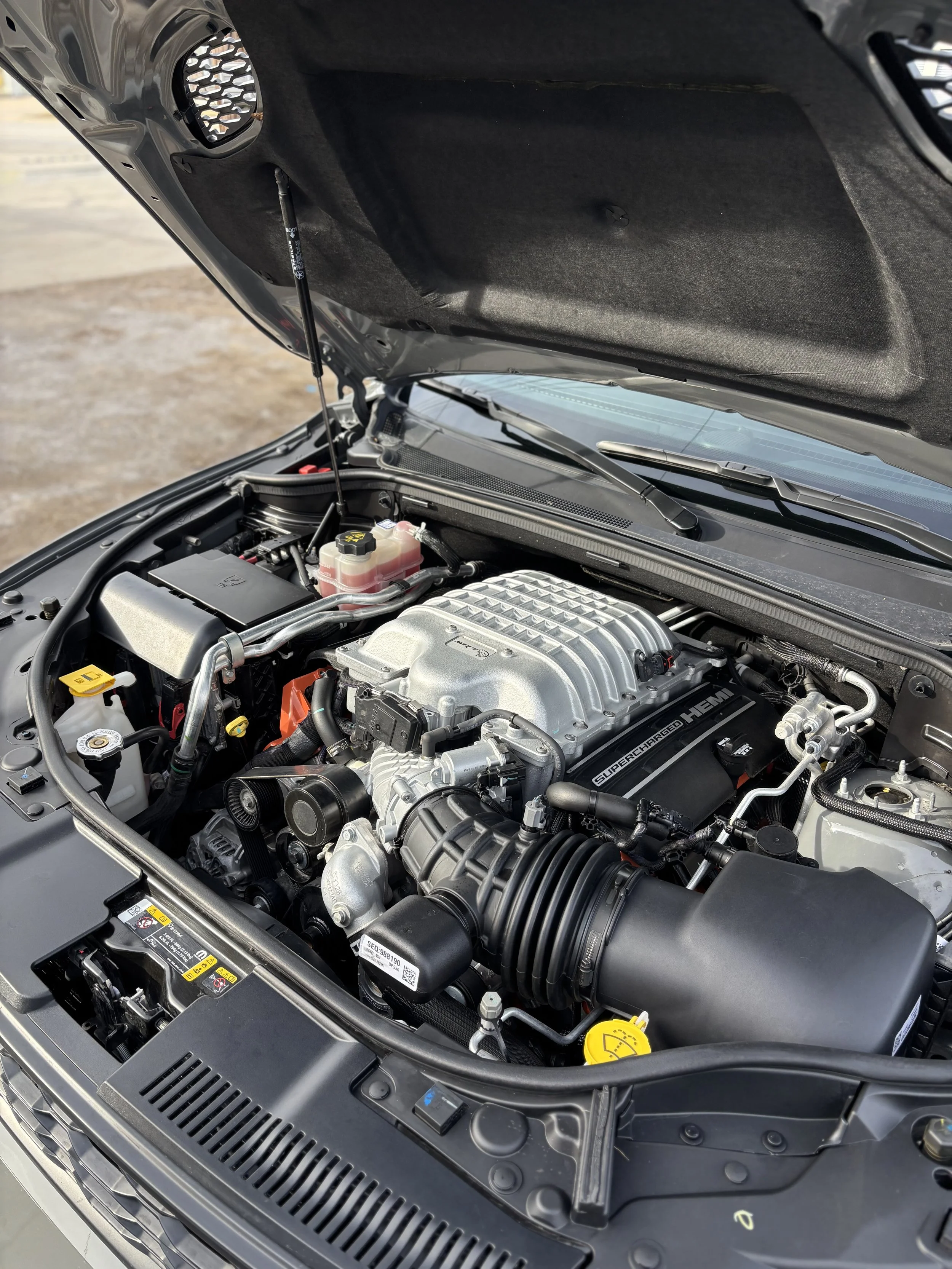 Close-up of a car engine bay showing a supercharged HEMI engine, various hoses, caps, and components, with the hood open.