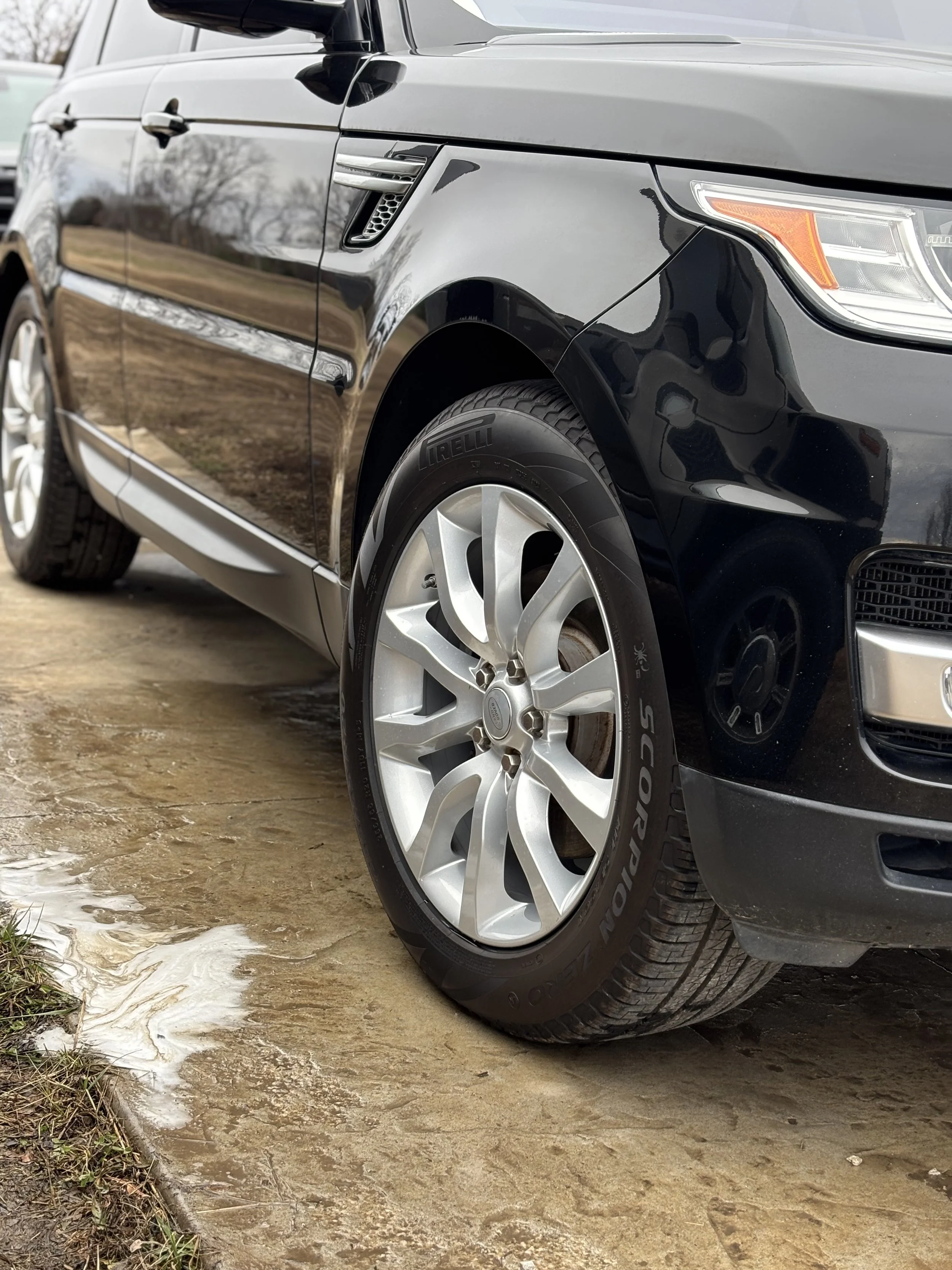 Close-up of the front right side of a black SUV showing a silver alloy wheel and a Pirelli tire, parked on a wet, dirt surface.