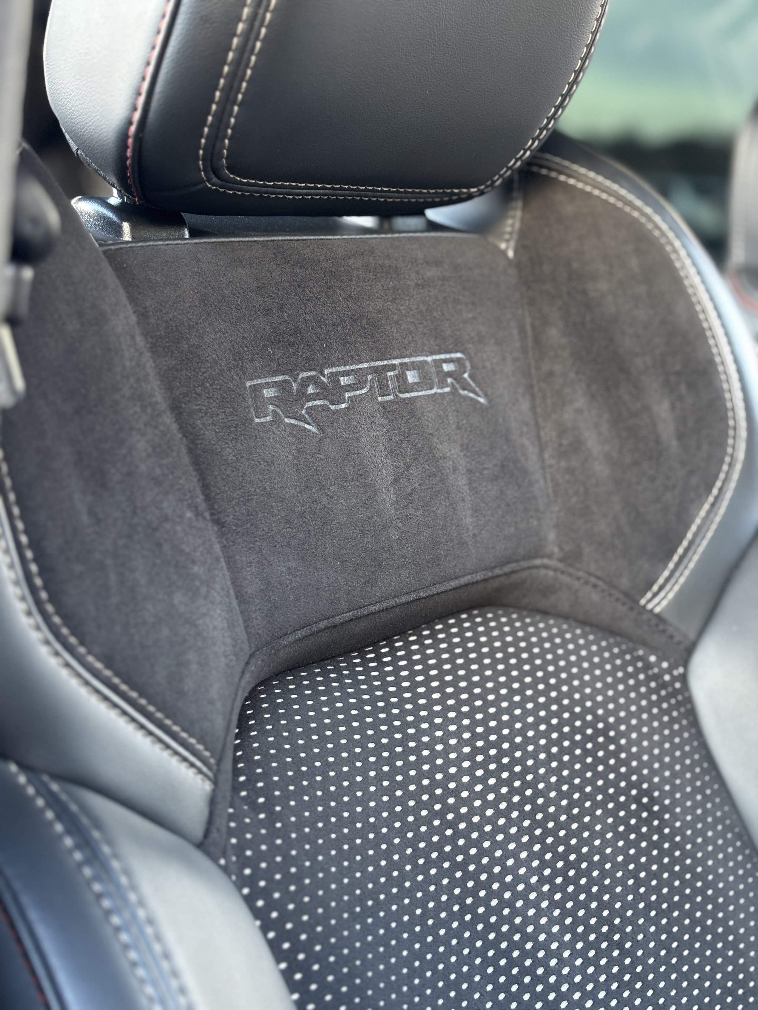 Close-up of a black car seat with the embroidered text 'Raptor' on the headrest area, featuring white contrast stitching and a dotted pattern on the lower part.