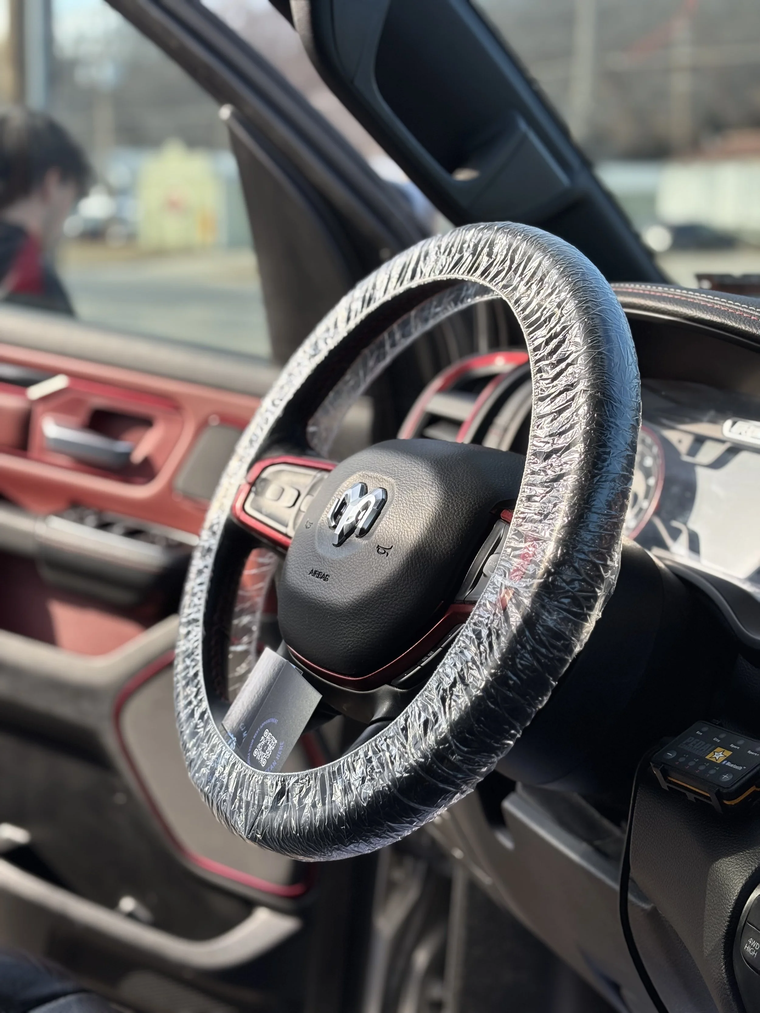 The interior of a vehicle showing a steering wheel wrapped in plastic for protection, with a dashboard and some controls visible.