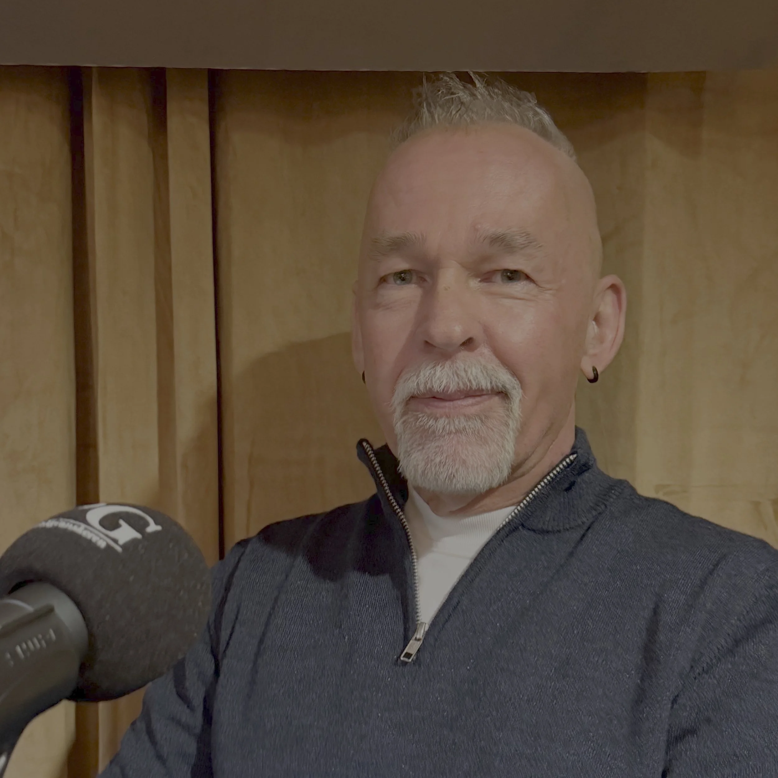 Person sitter på et podie foran et sceneteppe, iført mørk genser, med en mikrofon merket Privatgranskeren i forgrunnen under et podkastopptak.