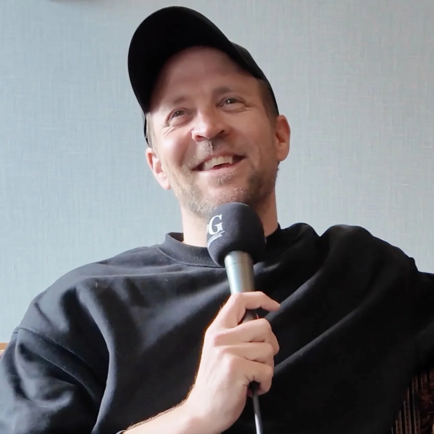 Person sitter i et podkastoppsett med mørk genser og caps, og holder en mikrofon med bokstaven «G» på. Bakgrunnen er en lys, ensfarget vegg med myk belysning.