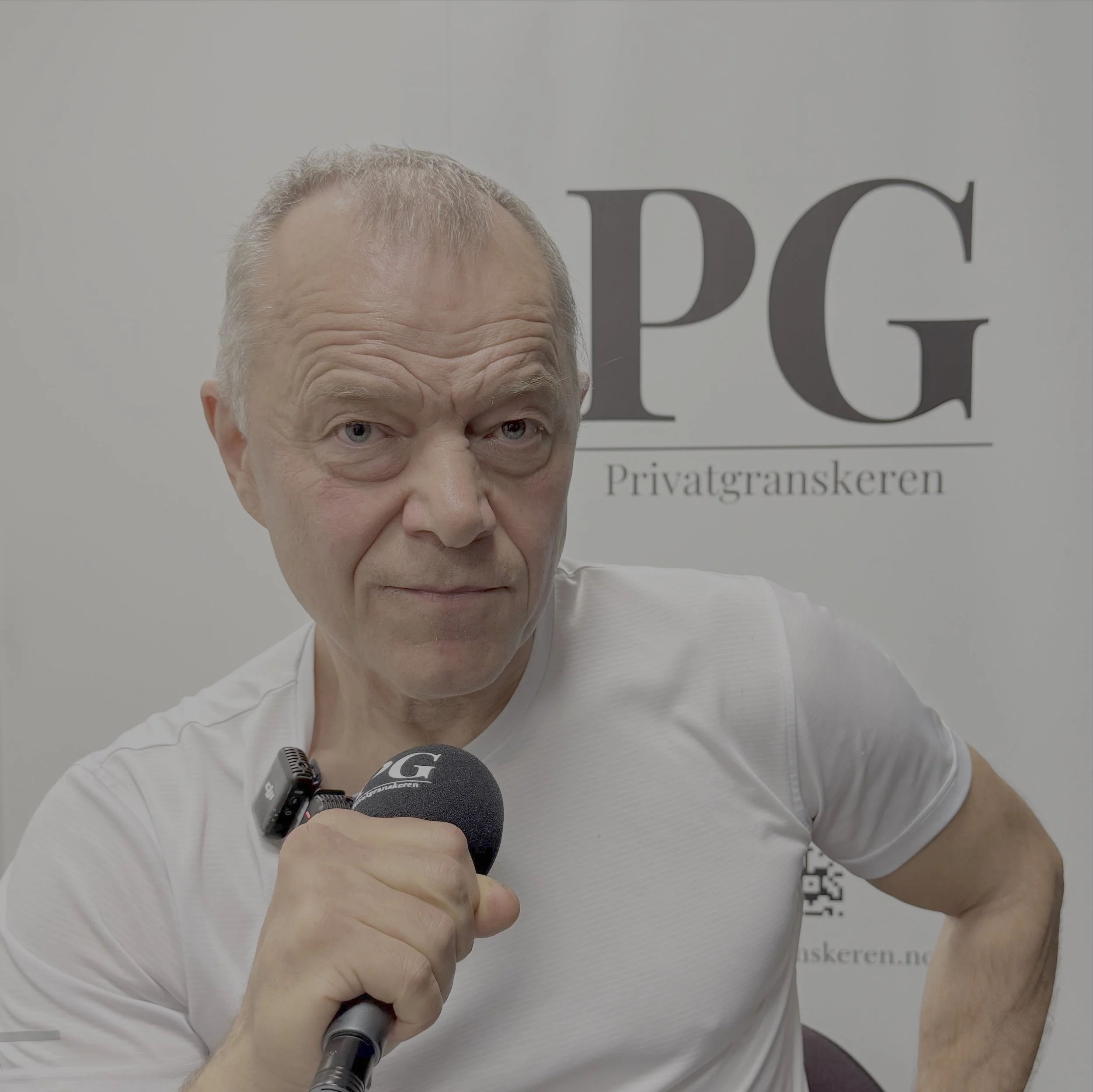 Person sitter i et podkaststudio med hvit t‑skjorte og holder en PG‑merket mikrofon. Bakgrunnen viser et lyst banner med store bokstaver fra Privatgranskeren‑logoen.