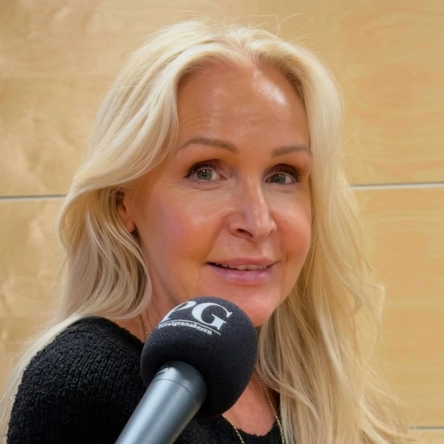 Person med langt lyst hår sitter i et podkastoppsett og holder en mikrofon med PG‑logo foran seg. Bakgrunnen består av lyse trepaneler med jevn belysning.