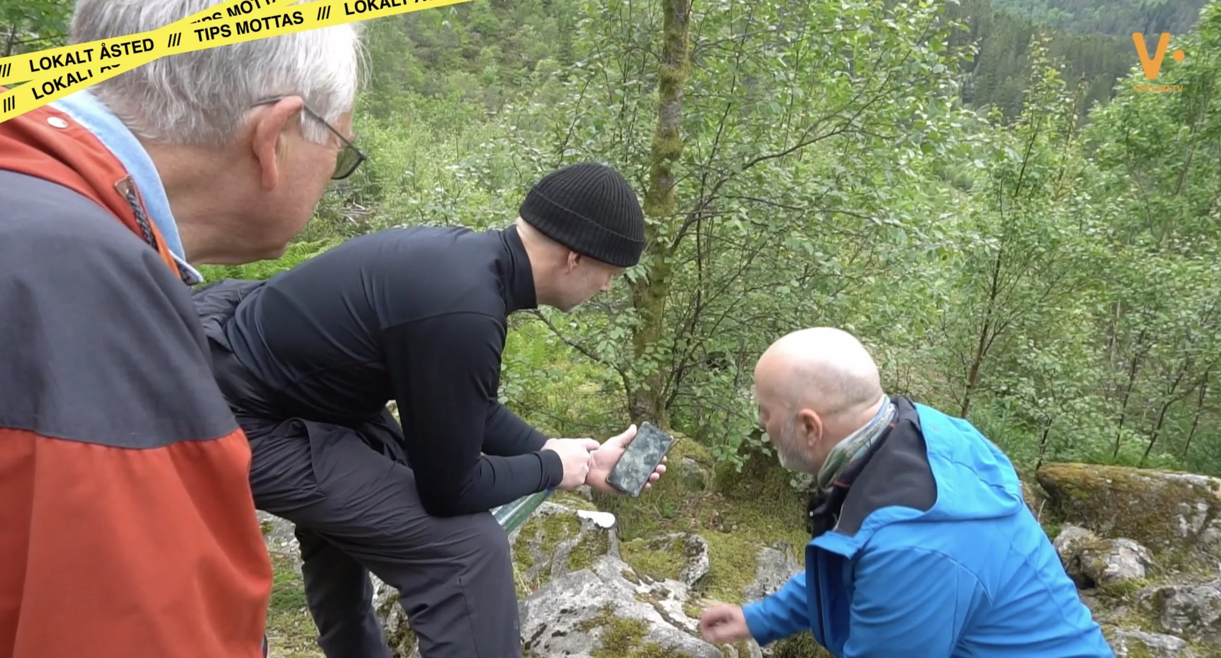 Flere personer undersøker et område i skogsterreng, der én person holder en mobiltelefon over steiner og mose. Gule bånd med teksten «LOKALT ÅSTED /// TIPS MOTTAS» strekker seg over toppen av bildet, og VSTLND.TV logoen vises i hjørnet.