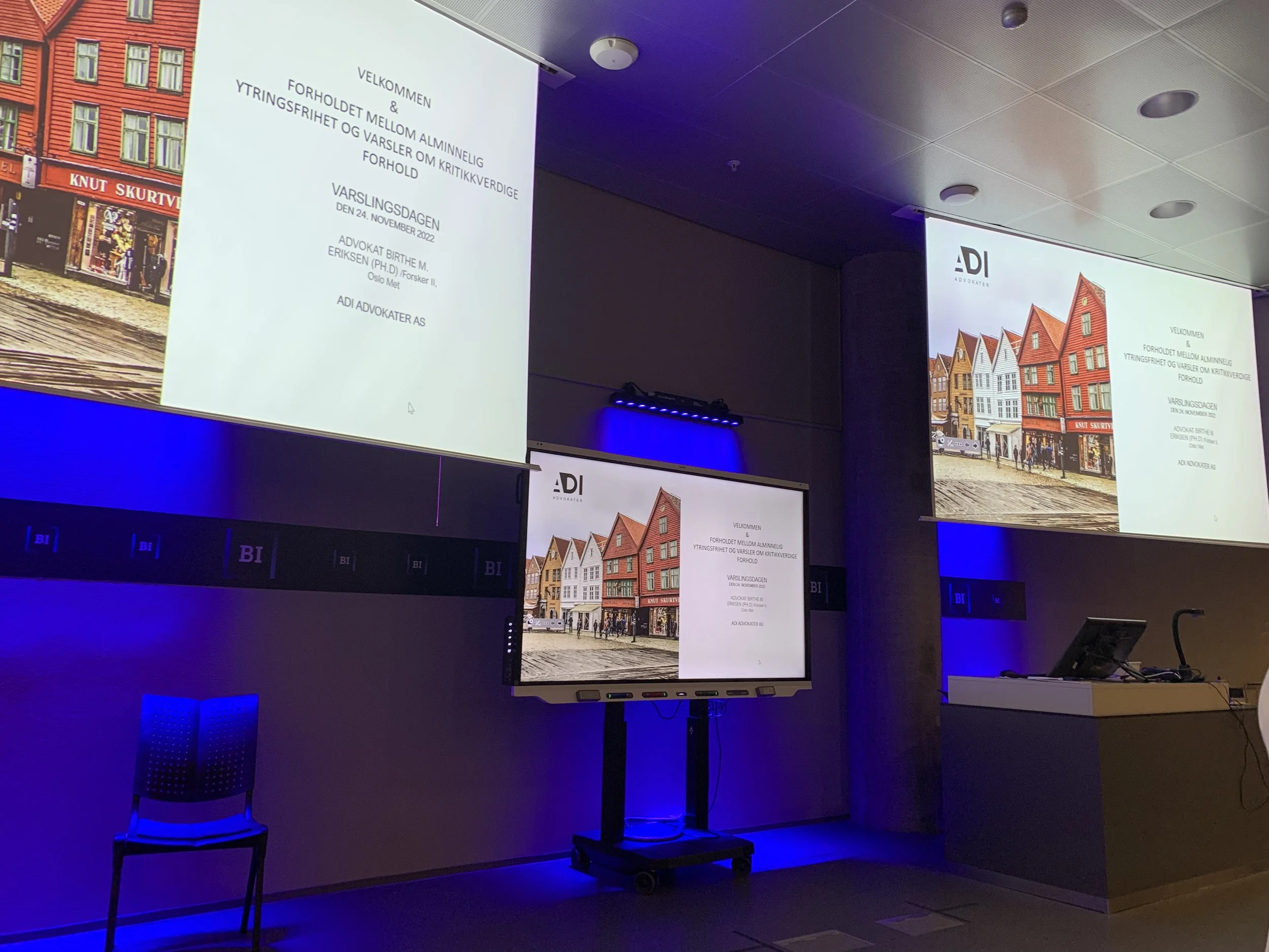 Foredragssal med tre store skjermer som viser presentasjonsslider med teksten «Velkommen» og tema om ytringsfrihet, varslere og rettslige forhold. Midt i rommet står en digital skjerm på stativ, og scenelyset er blått langs veggene.