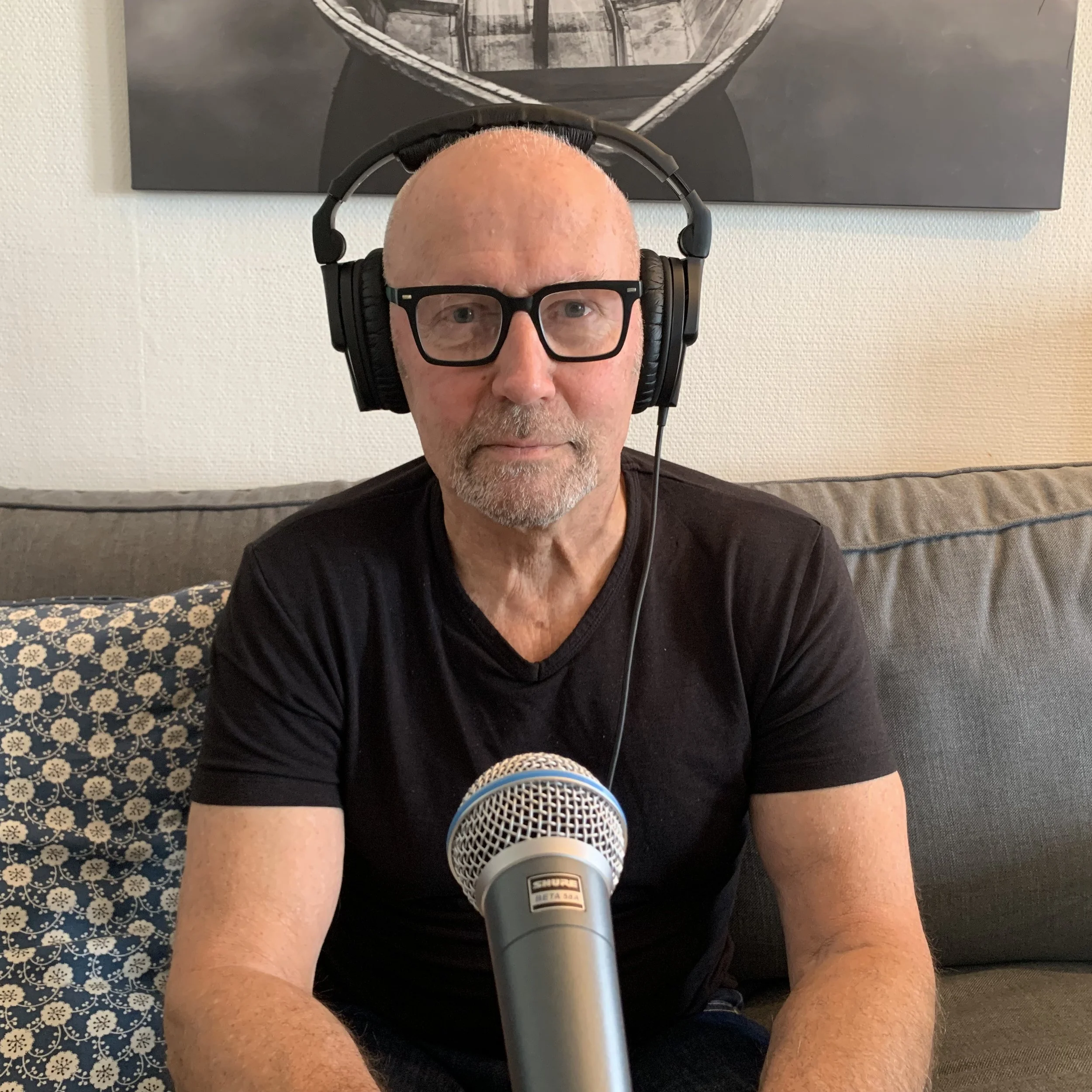 Person sitter på en sofa med hodetelefoner og snakker i en mikrofon, med et bilde på veggen bak og en pute med mønster ved siden av.