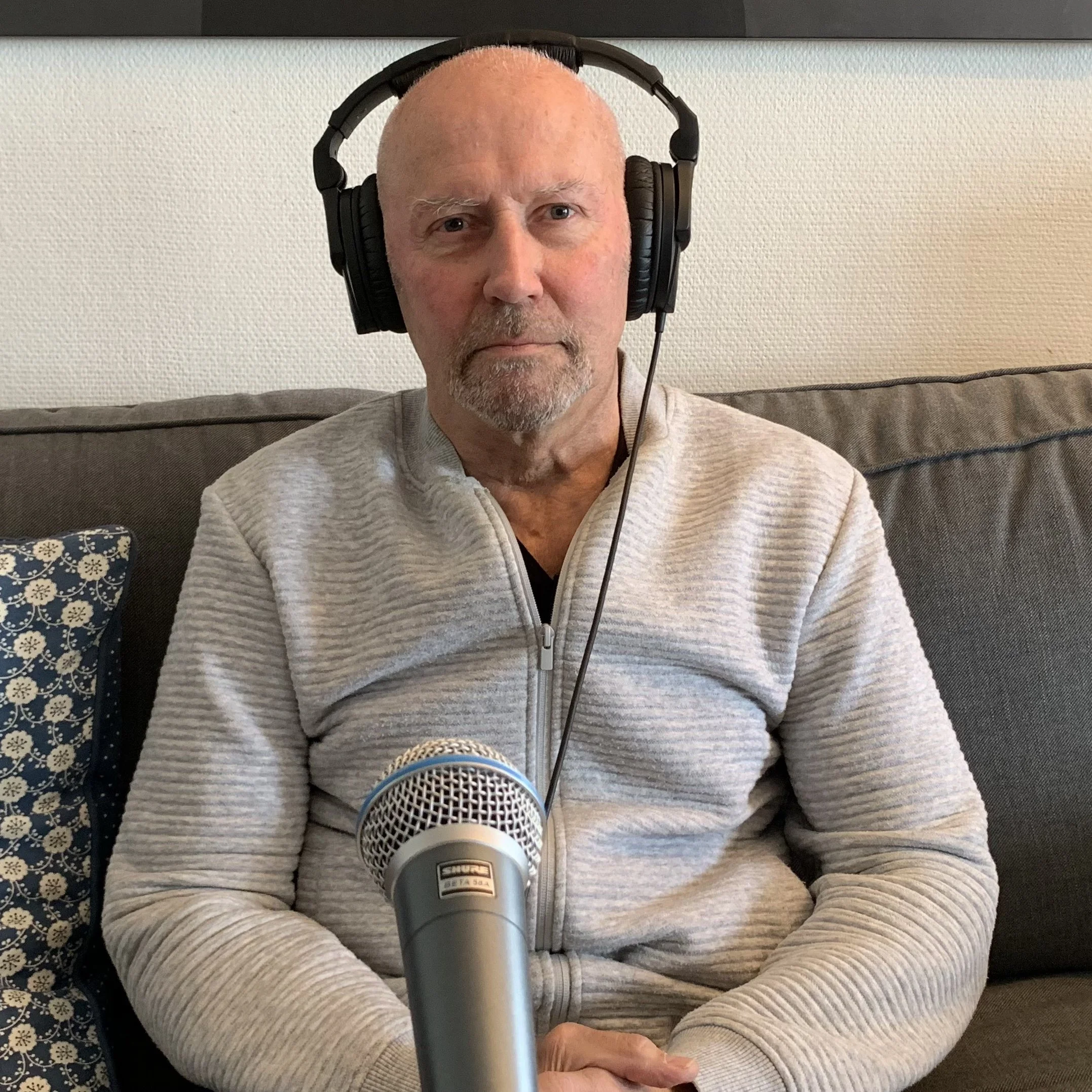 Person sitter på en sofa med hodetelefoner og en stor mikrofon plassert foran seg. Personen har på en lys, glidelåslukket jakke, og bakgrunnen viser en pute med mønster og en vegg med kunstmotiv.