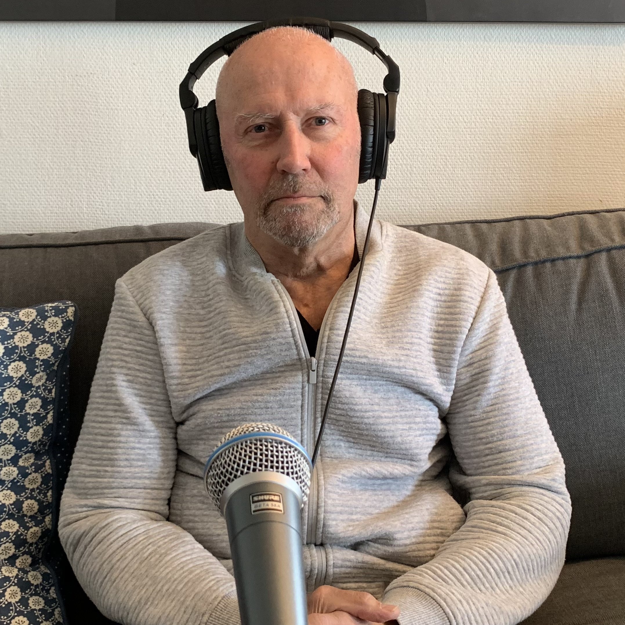 Person sitter på en sofa med hodetelefoner og en stor mikrofon plassert foran seg. Personen har på en lys, glidelåslukket jakke, og bakgrunnen viser en pute med mønster og en vegg med kunstmotiv.