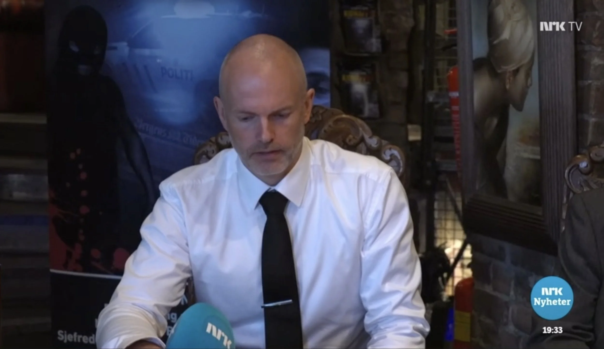 Person iført hvit skjorte og svart slips holder en NRK‑mikrofon under en pressekonferanse. I bakgrunnen sees en plakat med mørke motiv, en stol med utskårne detaljer samt mursteinsvegg og kunstverk. NRK Nyheter‑logoen vises nederst i høyre hjørne.