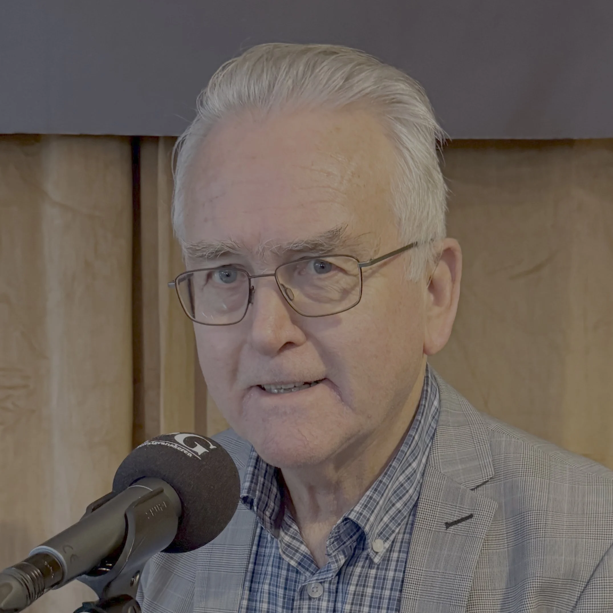 Person sitter i et podkastoppsett foran en PG‑merket mikrofon, iført rutet skjorte og lys blazer. Bakgrunnen består av lyse scenegardiner og en mørkere stoffplate øverst.