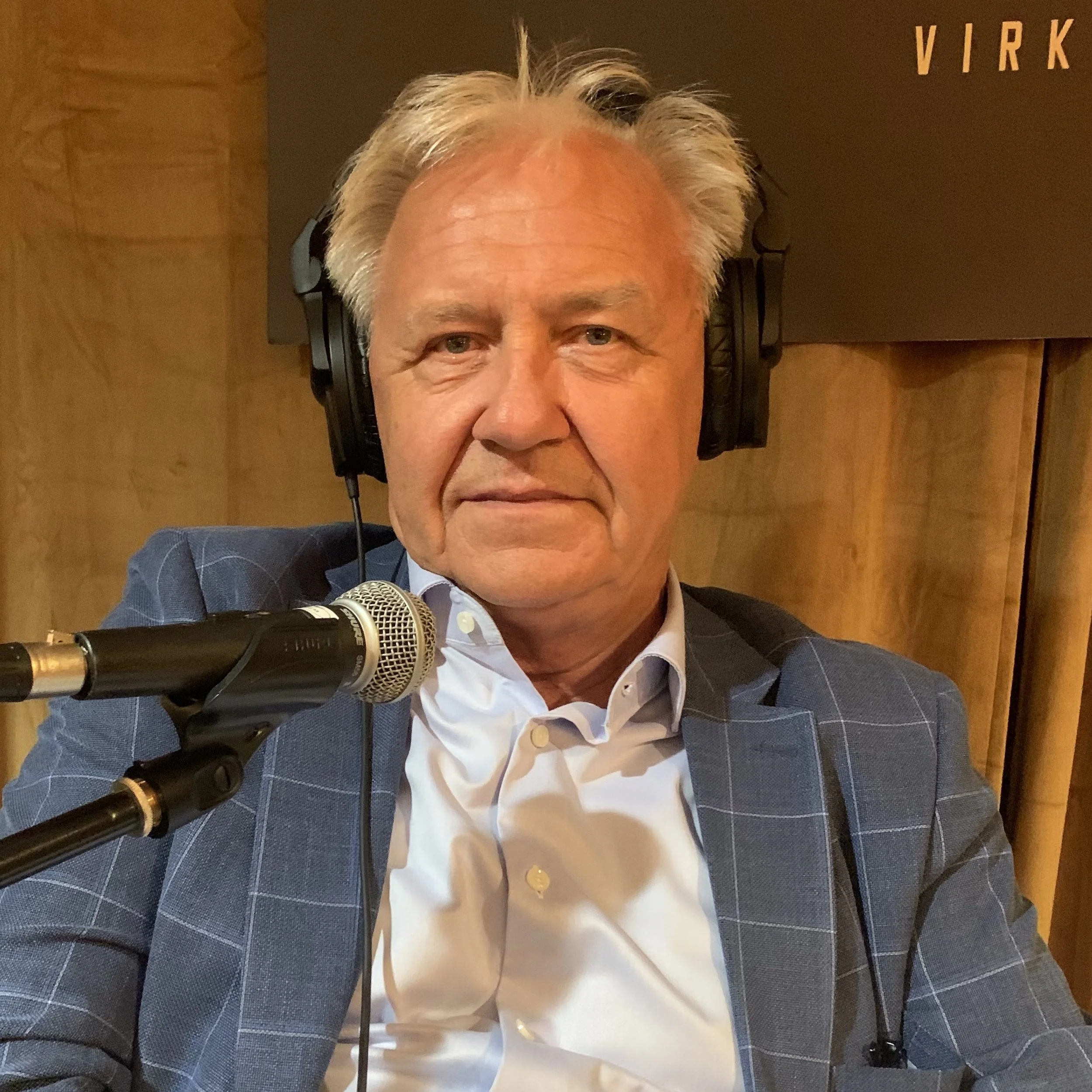 Person sitter i et podkaststudio med hodetelefoner og mikrofon foran seg, iført en rutete blazer og lys skjorte. Bakgrunnen består av et brunt sceneteppe og en mørk bannervegg.