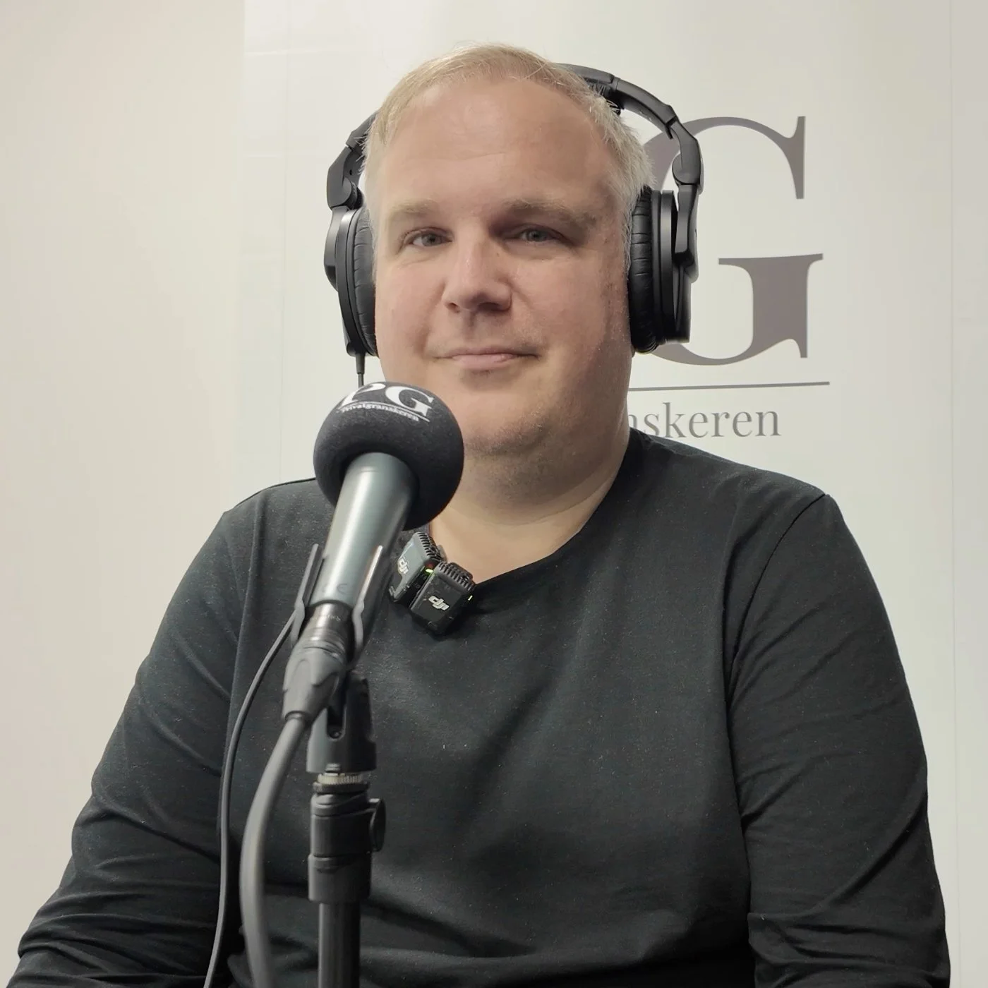 Person i podkaststudio bruker store hodetelefoner og snakker i en mikrofon merket «PG». I bakgrunnen vises en lys vegg med deler av logoen til Privatgranskeren.