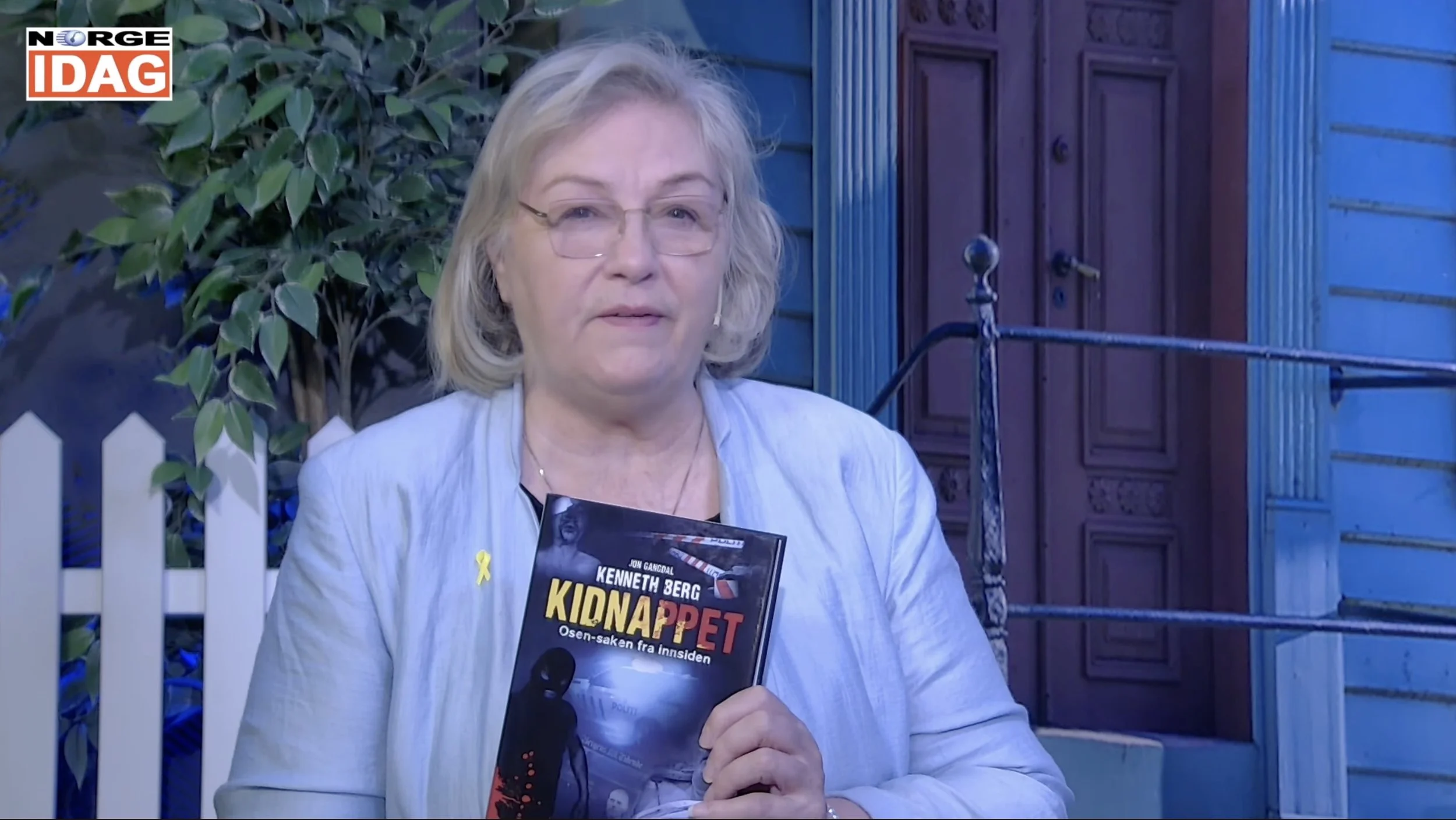 Person i lyst antrekk holder frem boken «Kidnappet – Osen‑saken fra innsiden» i et studiooppsett. I bakgrunnen ses et hvitt hagegjerde, grønne planter og en blå trehusfasade med dør og vindu. Norge IDAG‑logoen vises øverst i hjørnet.