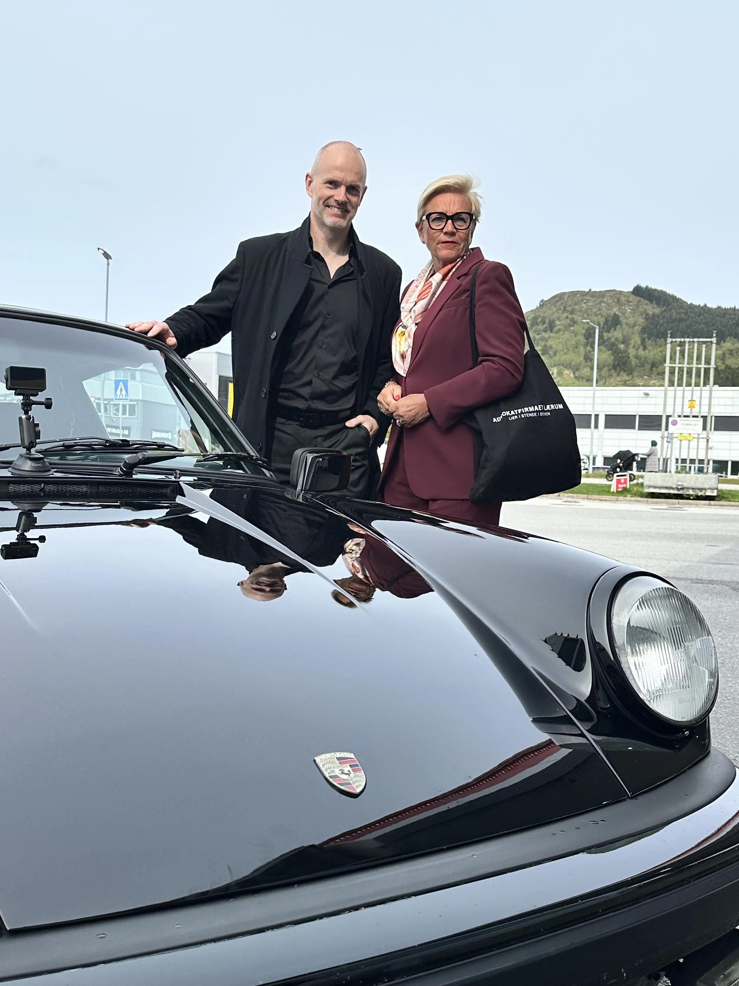 To personer står ved en svart klassisk Porsche 911 parkert utendørs, med bygninger og fjell i bakgrunnen.