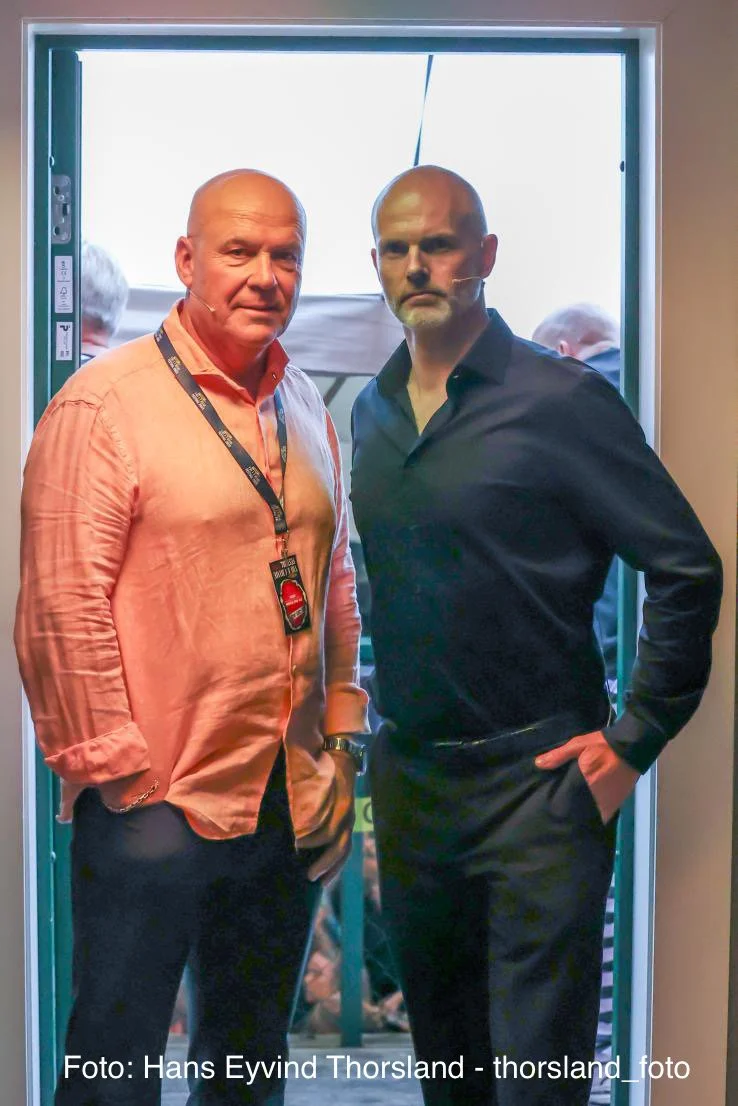 To personer står side om side i en døråpning på et arrangement, én i en lys skjorte med festivalpass rundt halsen og én i en mørk skjorte. Bakgrunnen viser folk i et uteområde.