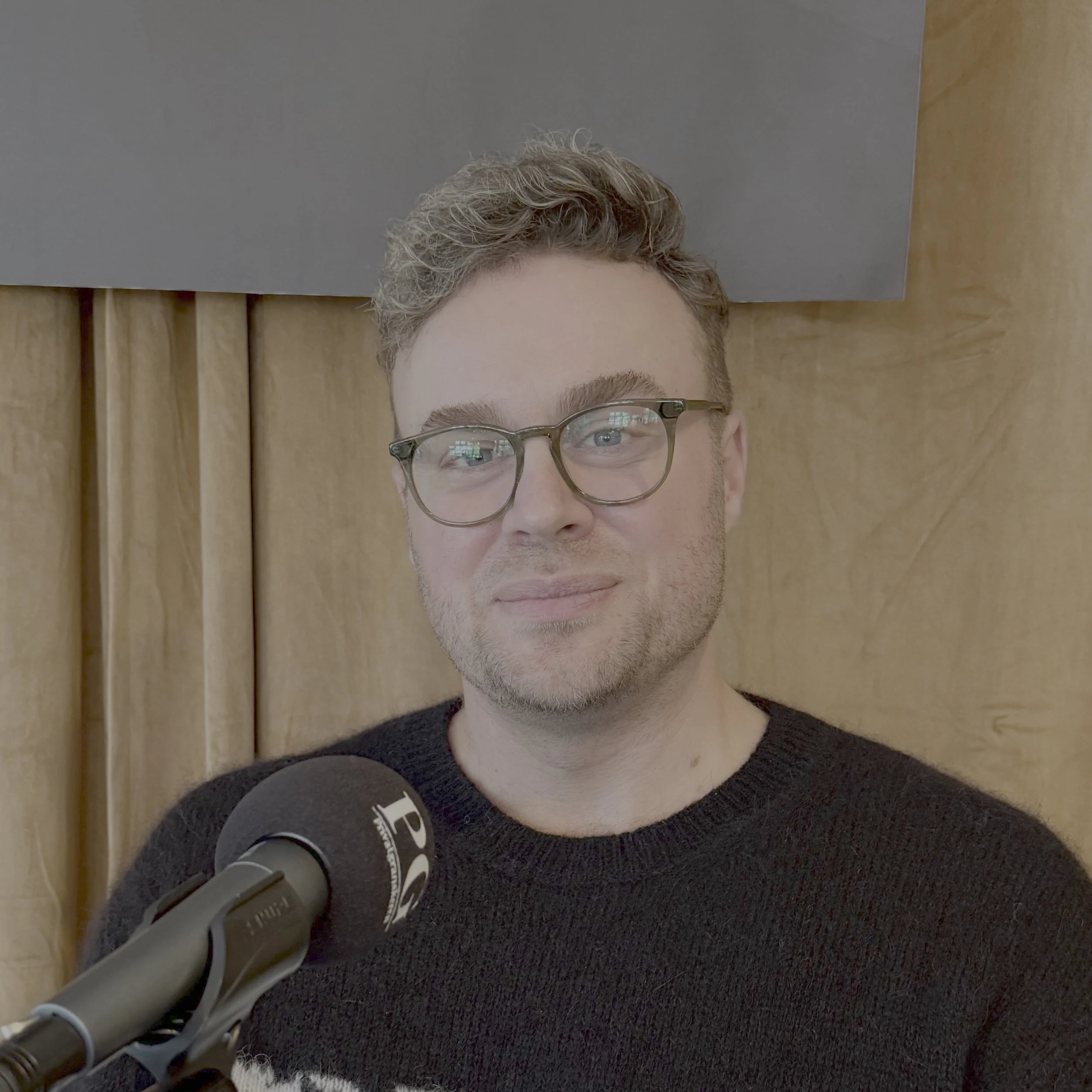 Person sitter i et podkastoppsett foran en mikrofon merket «PG». Personen har kort, bølget hår øverst og er kledd i en mørk genser. Bakgrunnen består av et lyst sceneteppe i tekstil.