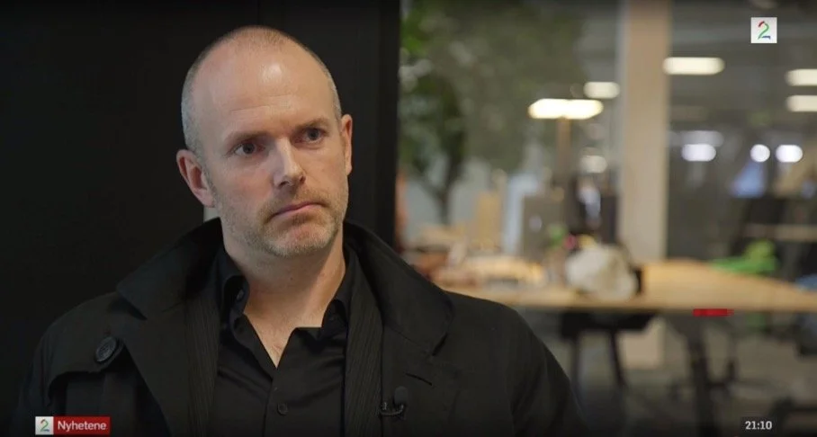 Person i mørk frakk og mørk skjorte sitter i et TV studio under et intervju. I bakgrunnen vises et kontorlandskap med skrivebord, dataskjermer, lamper og glassflater. TV 2 logoen er plassert øverst til høyre i bildet, og et nyhetsbanner nederst.