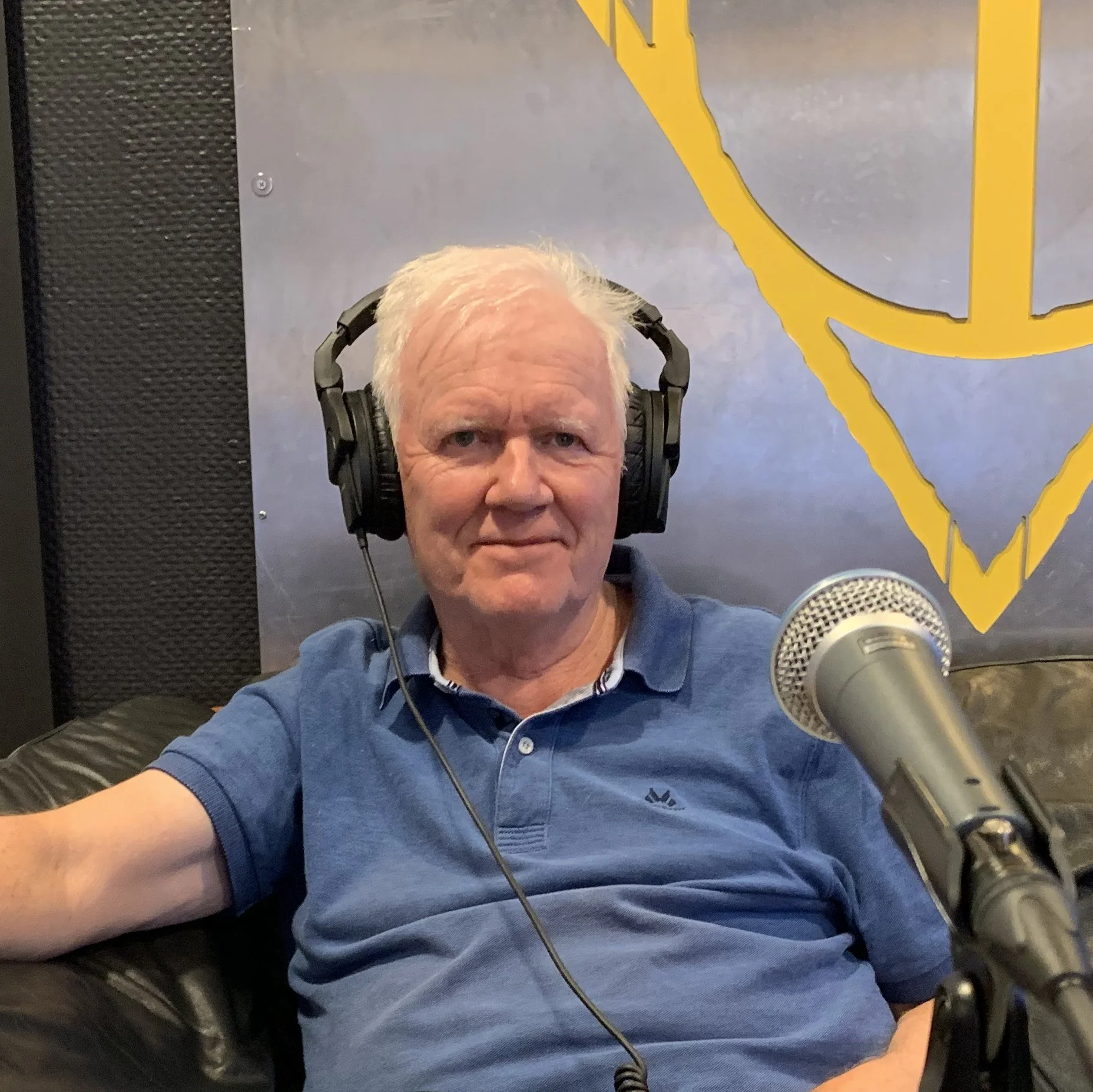 Person i et podcaststudio sitter i en stol med hodetelefoner foran en mikrofon. Bakgrunnen viser lyddempende veggpaneler og et veggmaleri i gule linjer.