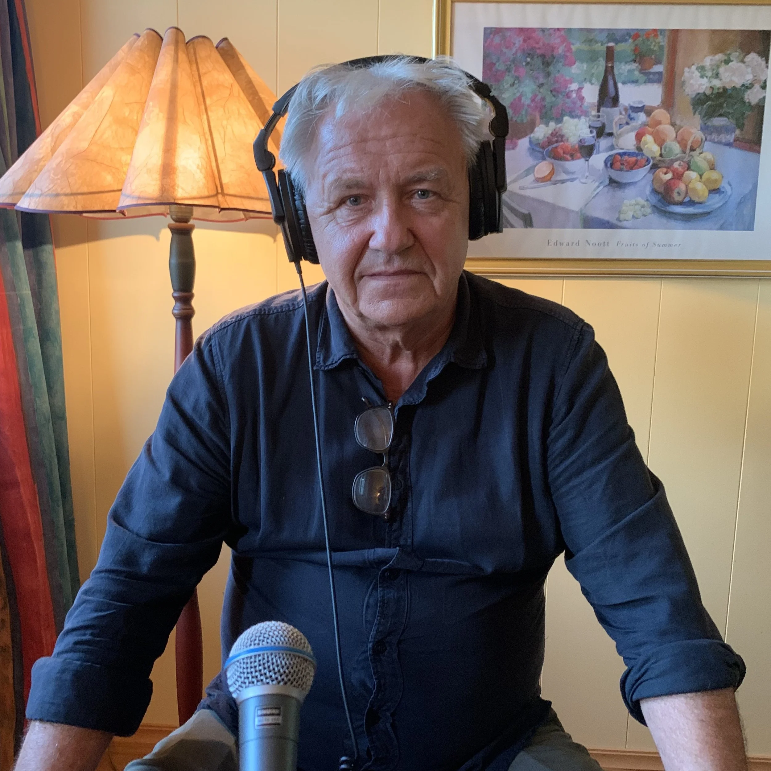 Person sitter i et hjemmestudio med hodetelefoner og mikrofon plassert foran seg. Personen har på en mørk skjorte, og et par briller henger i skjortekanten. Bakgrunnen viser en gul vegg med et innrammet maleri, samt en gulvlampe med stoffskjerm.