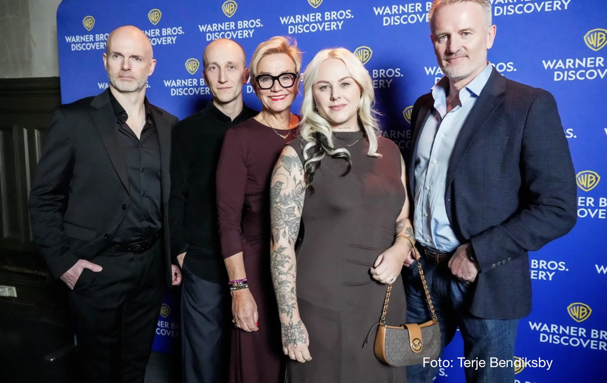 Fem personer står samlet foran en blå pressevegg med gjentatte logoer for Warner Bros. Discovery. Personene er kledd i formelle antrekk som dressjakker, skjorter og kjoler.