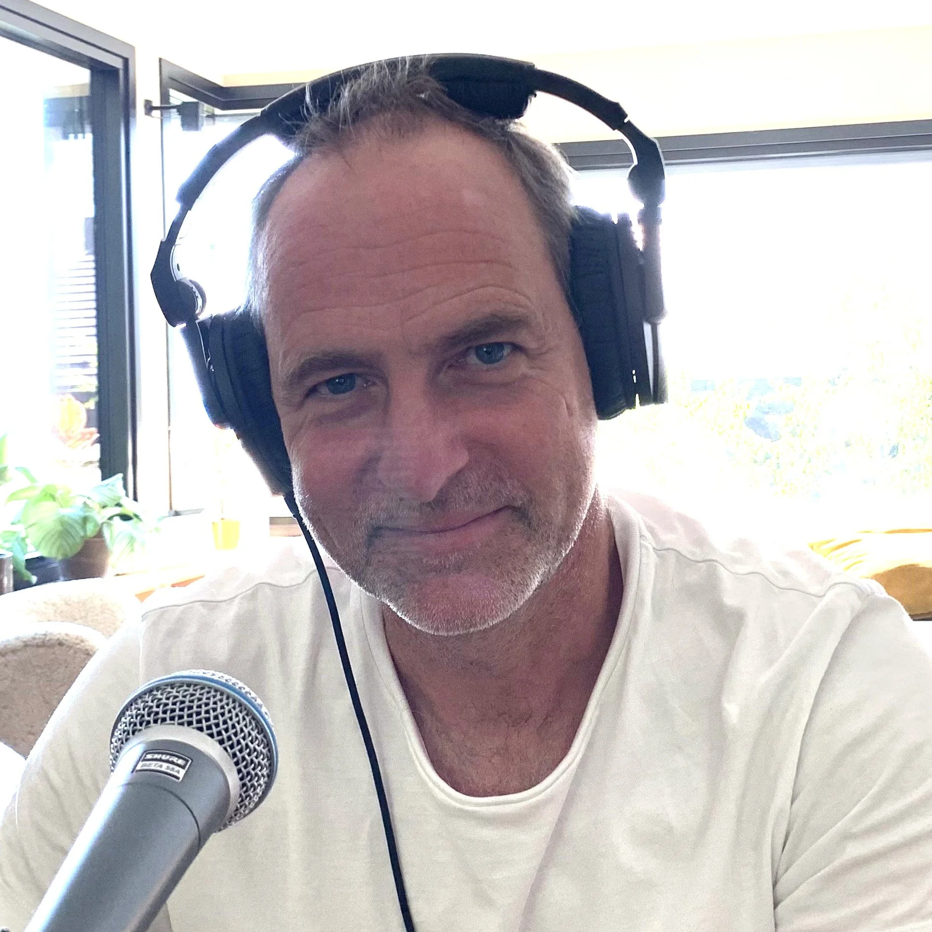 Person sitter innendørs foran en mikrofon med hodetelefoner på, iført en lys T‑skjorte. I bakgrunnen er det store vinduer, planter og et lyst interiør.