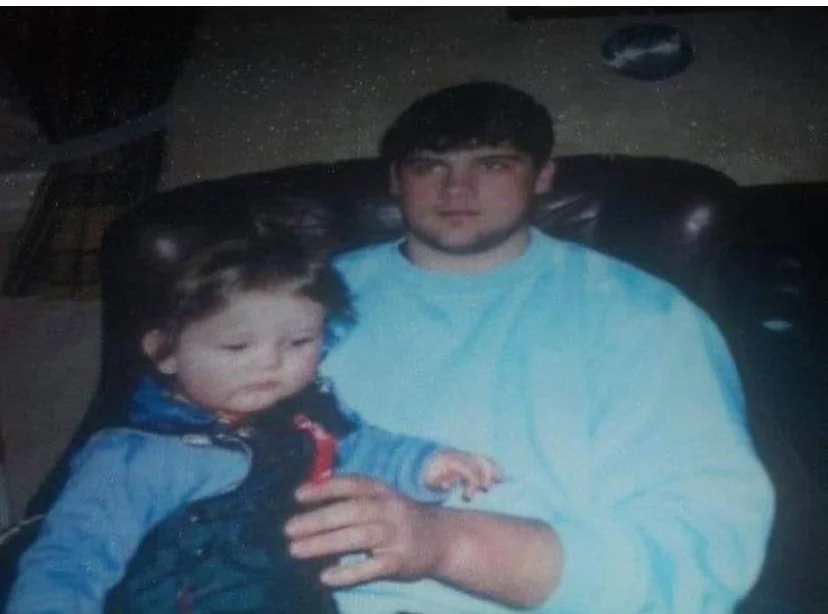 To personer sitter sammen i en mørk skinnstol. Den ene holder et lite barn i fanget, som er kledd i blå yttertøy. Bakgrunnen viser en vegg med en tallerken på som pynt.