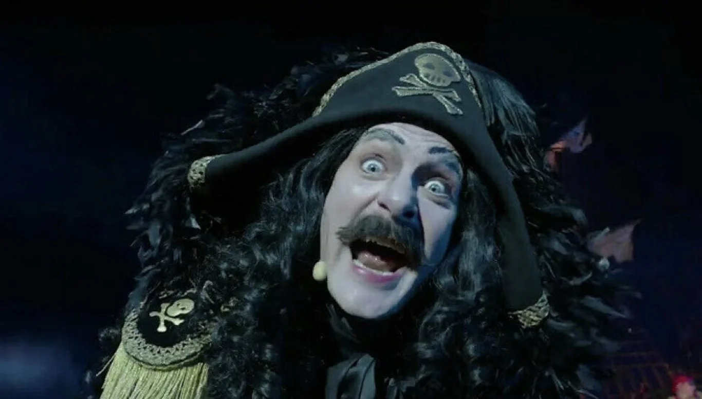 En person kledd i et dramatisk svart piratinspirert kostyme med fjær og hodeplagg med hodeskallemotiv, opplyst i et mørkt scenemiljø.