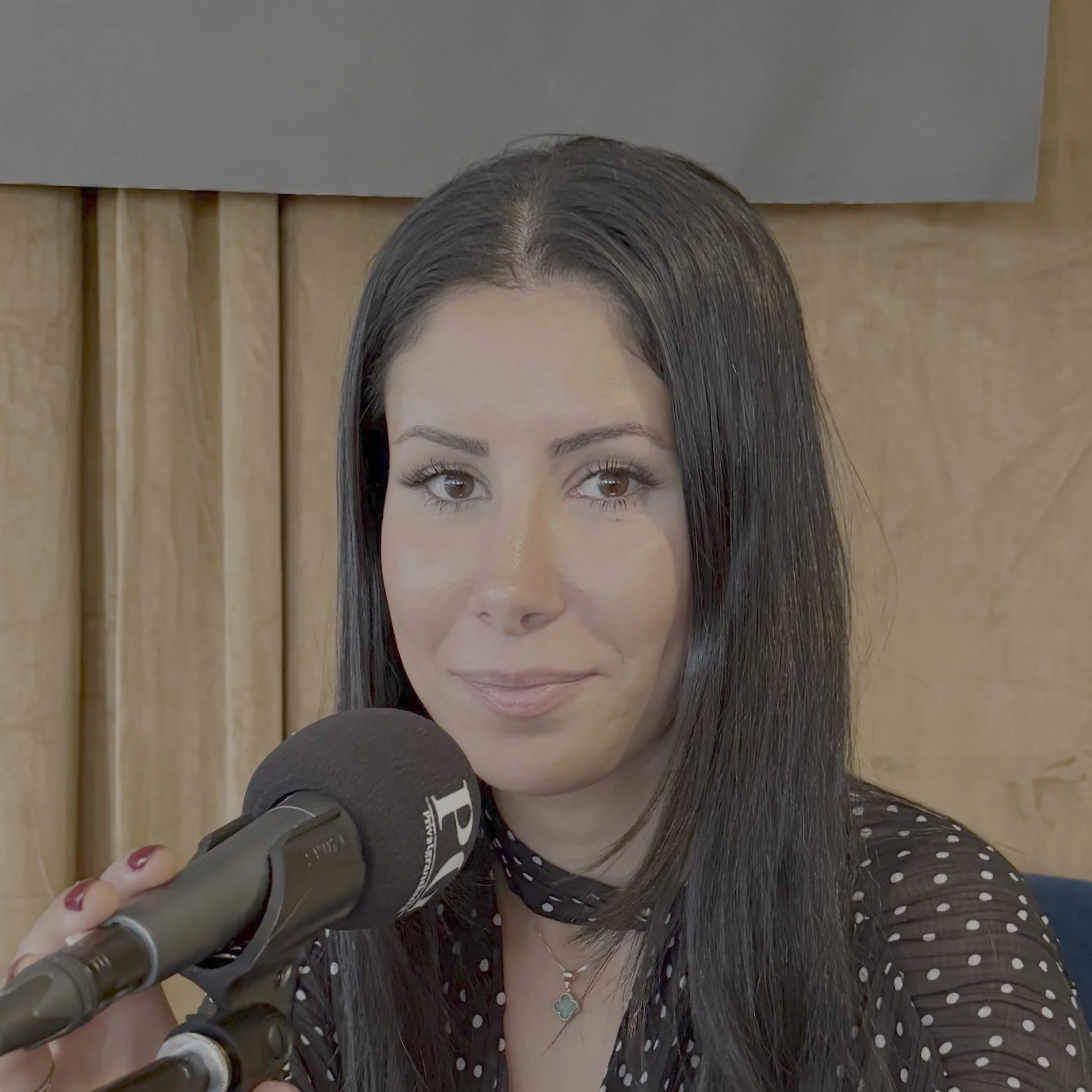 Person med langt, mørkt hår sitter i et podkastoppsett og holder en PG‑merket mikrofon. Personen er kledd i en mørk bluse og har et smalt smykke rundt halsen. Bakgrunnen består av lyse scenegardiner og en mørk panelplate øverst i bildet.