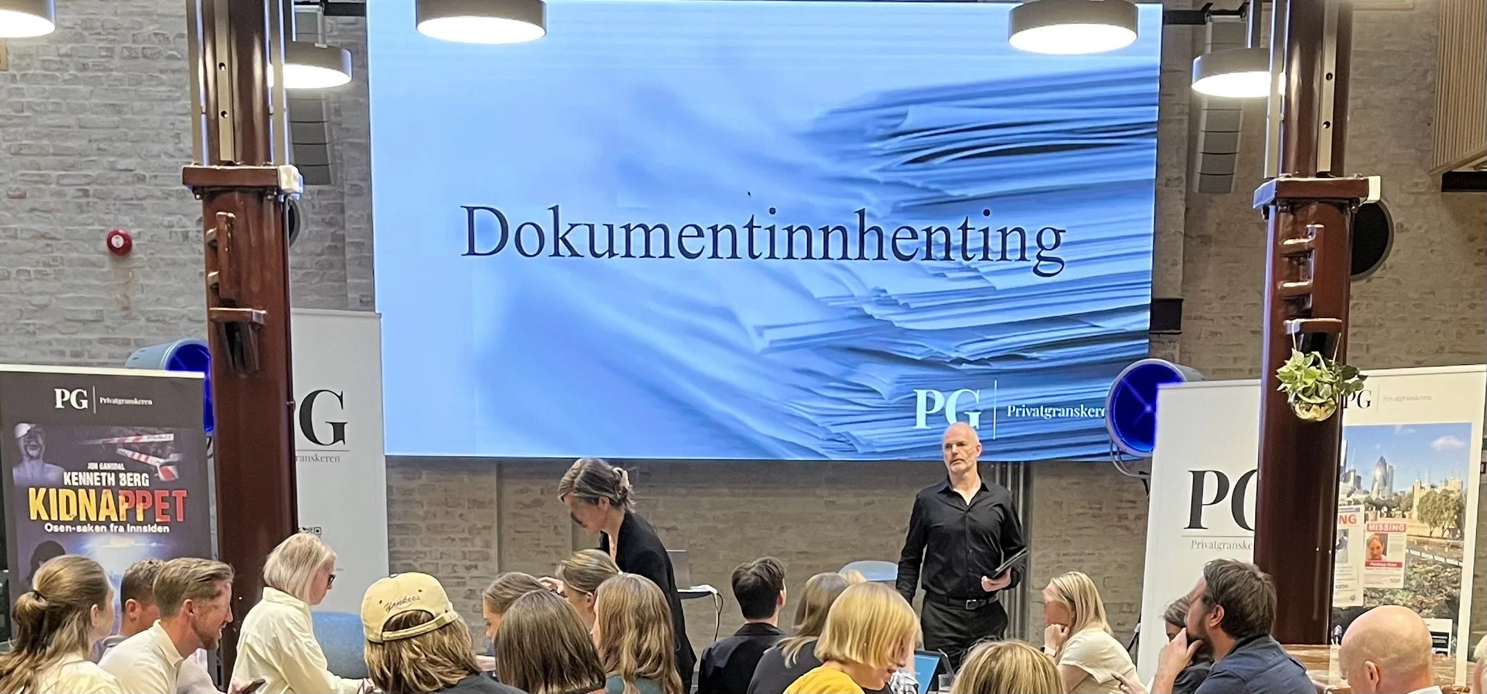 Foredrag i en sal med en stor skjerm som viser teksten «Dokumentinnhenting» over et bilde av stabler med dokumenter. Publikum sitter ved bord foran scenen, og to personer står ved presentasjonsområdet.