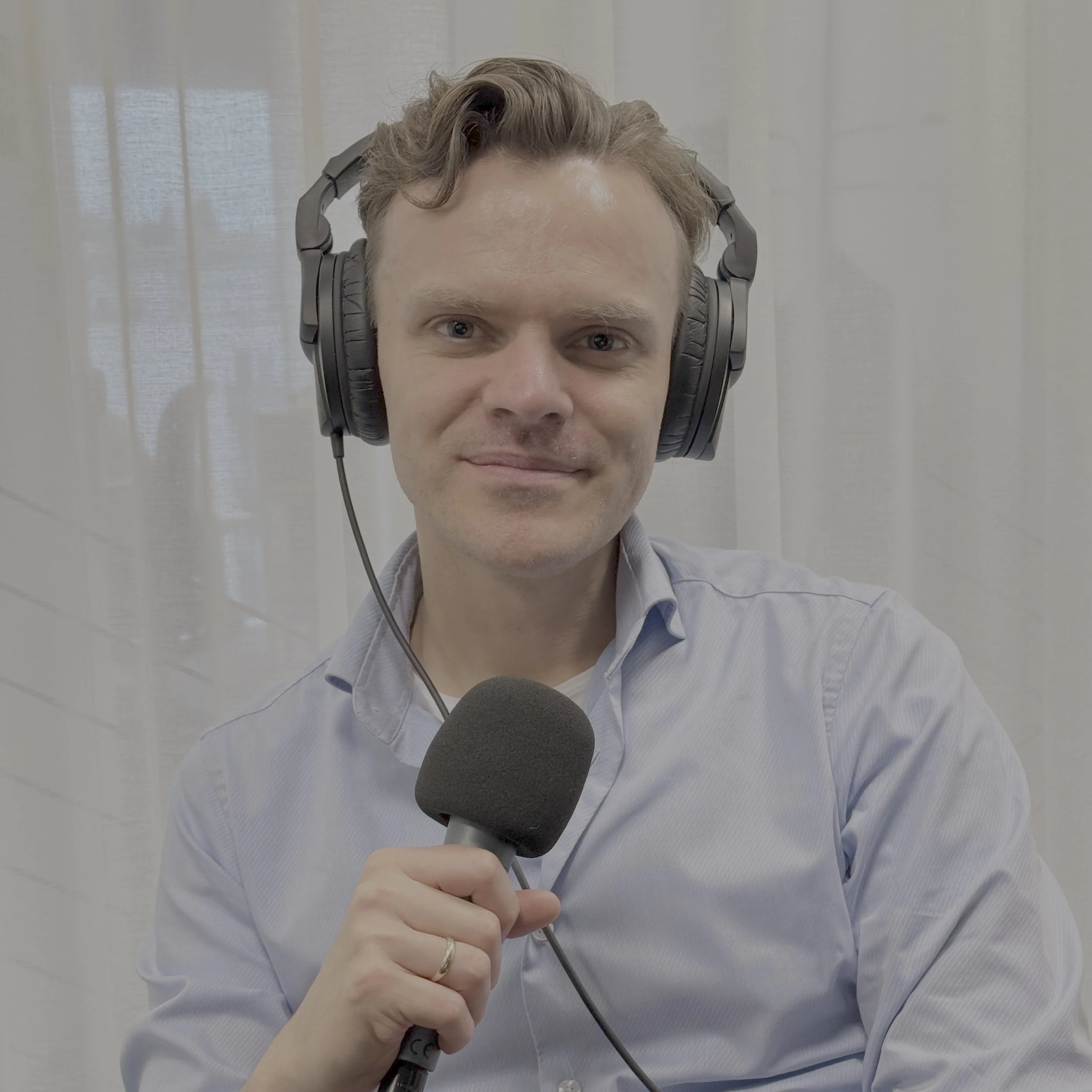 Person sitter i et podkastopptak med store hodetelefoner og holder en håndholdt mikrofon. Personen har på en lys blå skjorte, og bakgrunnen består av lyse gardiner med mildt filtrert dagslys.