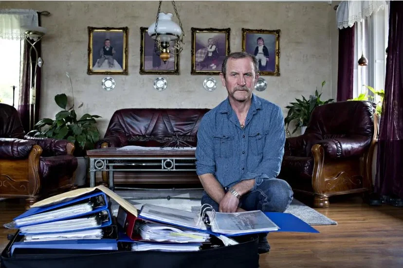 Person sitter på gulvet i en stue omgitt av flere åpne permer og dokumentmapper. Rommet har mørke skinnmøbler, trepanel på gulvet, grønne planter og fire innrammede portrettbilder på veggen bak.